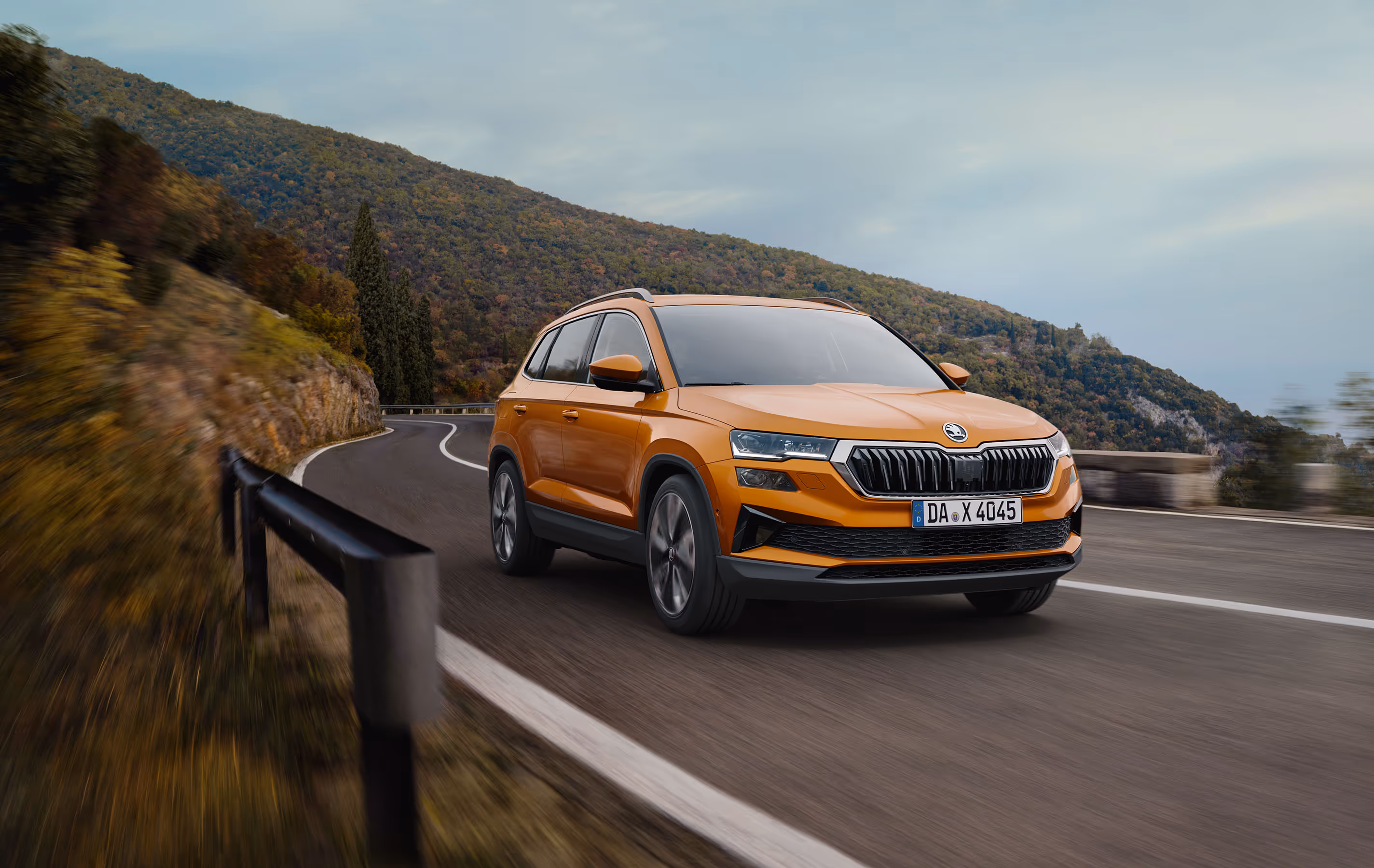 Ein Škoda Karoq in der Lackierung Phoenix-Orange Metallic fährt auf einer kurvenreichen Straße entlang einer Felswand. Das Fahrzeug wird in einer dynamischen Dreiviertelansicht von vorne gezeigt, während die Bewegungsunschärfe des Hintergrunds die Fahrszene unterstreicht. Unten rechts steht auf einer dunkelgrün abgesetzten Fläche "Sicherheit & Assistenten" geschrieben.