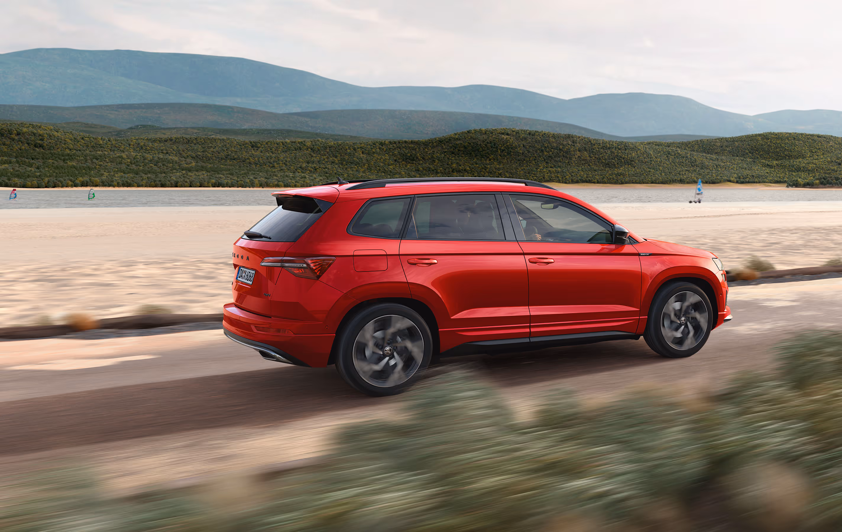 Ein Škoda Karoq Sportline in der Lackierung Velvet-Rot Metallic fährt auf einer Straße entlang einer Küstenlandschaft. Die Aufnahme von schräg hinten zeigt die dynamische Fahrszene, unterstrichen durch Bewegungsunschärfe, und hebt die schwarzen Design-Details wie den Heckspoiler und die Dachreling hervor. Unten rechts steht auf einer dunkelgrün abgesetzten Fläche "Sportline" geschrieben.