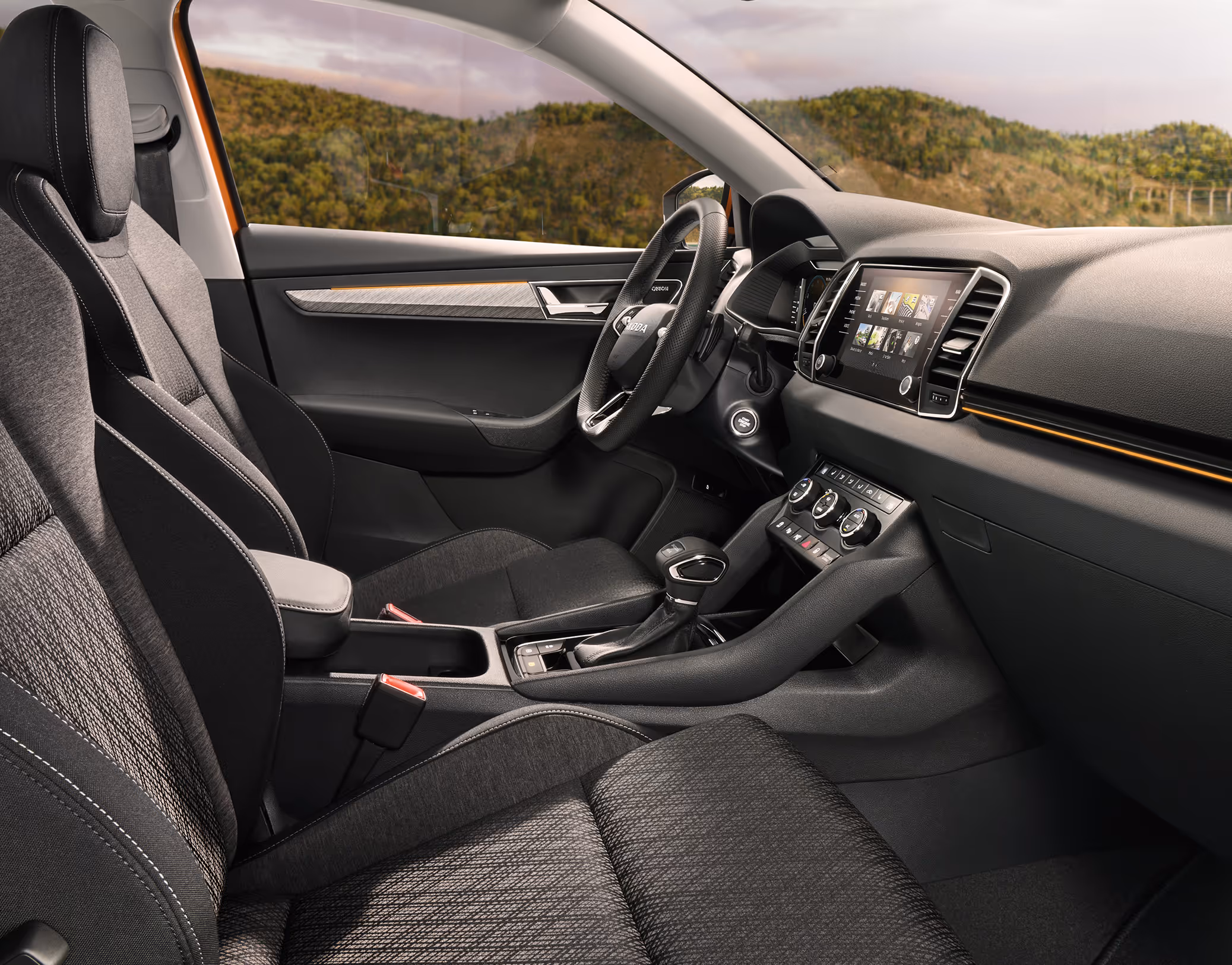 Eine seitliche Innenansicht des Škoda Karoq zeigt das Interieur der Design Selection Lodge. Die Sitzbezüge aus einer Stoff-Suedia-Kombination, die durch kupferfarbene Kontrastnähte akzentuiert werden, sowie das dunkle Armaturenbrett mit kupferfarbener Dekorleiste sind zu sehen, während durch die Fenster eine hügelige, bewaldete Landschaft sichtbar ist.