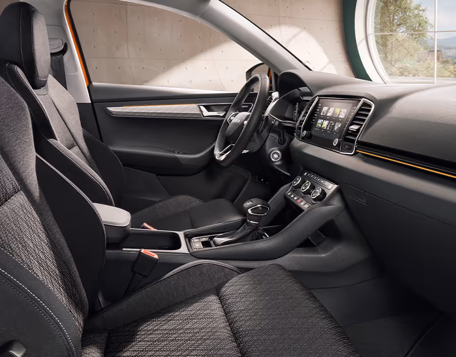 Eine seitliche Innenansicht des Škoda Karoq zeigt das Interieur der Design Selection Lodge. Die Sitzbezüge aus einer Stoff-Suedia-Kombination, die durch kupferfarbene Kontrastnähte akzentuiert werden, sowie das dunkle Armaturenbrett mit kupferfarbener Dekorleiste sind zu sehen, während durch die Fenster eine hügelige, bewaldete Landschaft sichtbar ist.