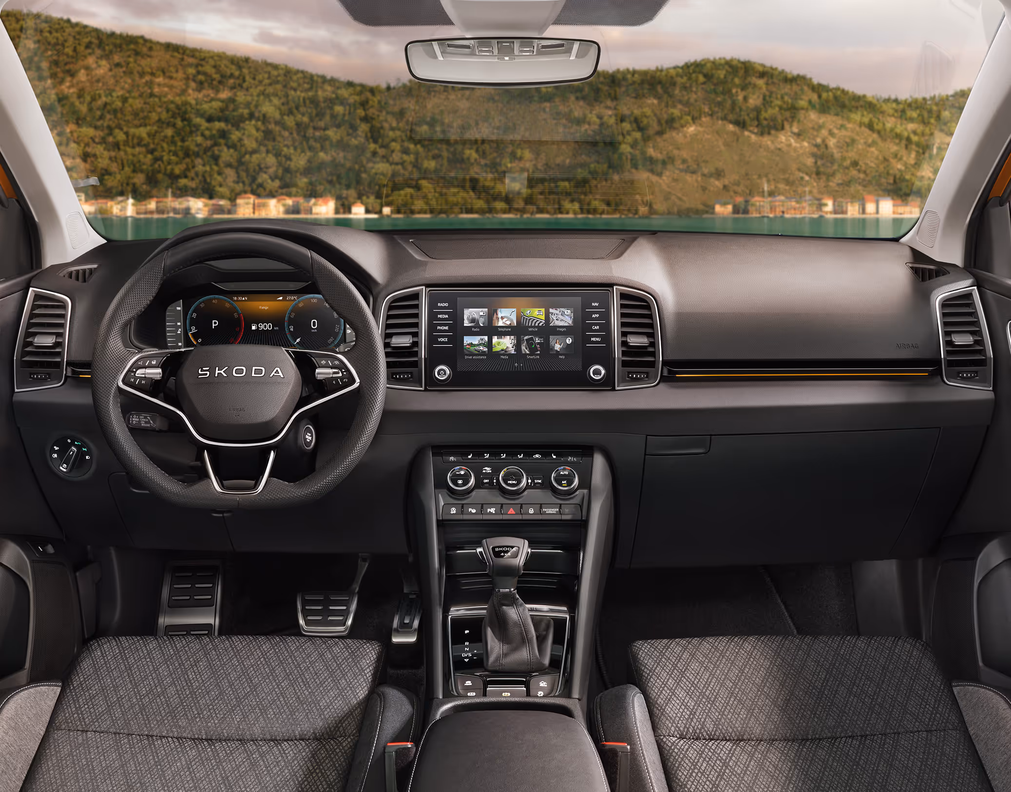 Eine weite Innenansicht zeigt das gesamte Cockpit eines Škoda Karoq mit der Design Selection Lodge. Das dunkle Interieur ist mit Sitzbezügen aus einer Stoff-Suedia-Kombination und einer kupferfarbenen Dekorleiste am Armaturenbrett ausgestattet. Durch die Windschutzscheibe ist eine Küstenlandschaft mit Hügeln und einem Gewässer zu sehen.