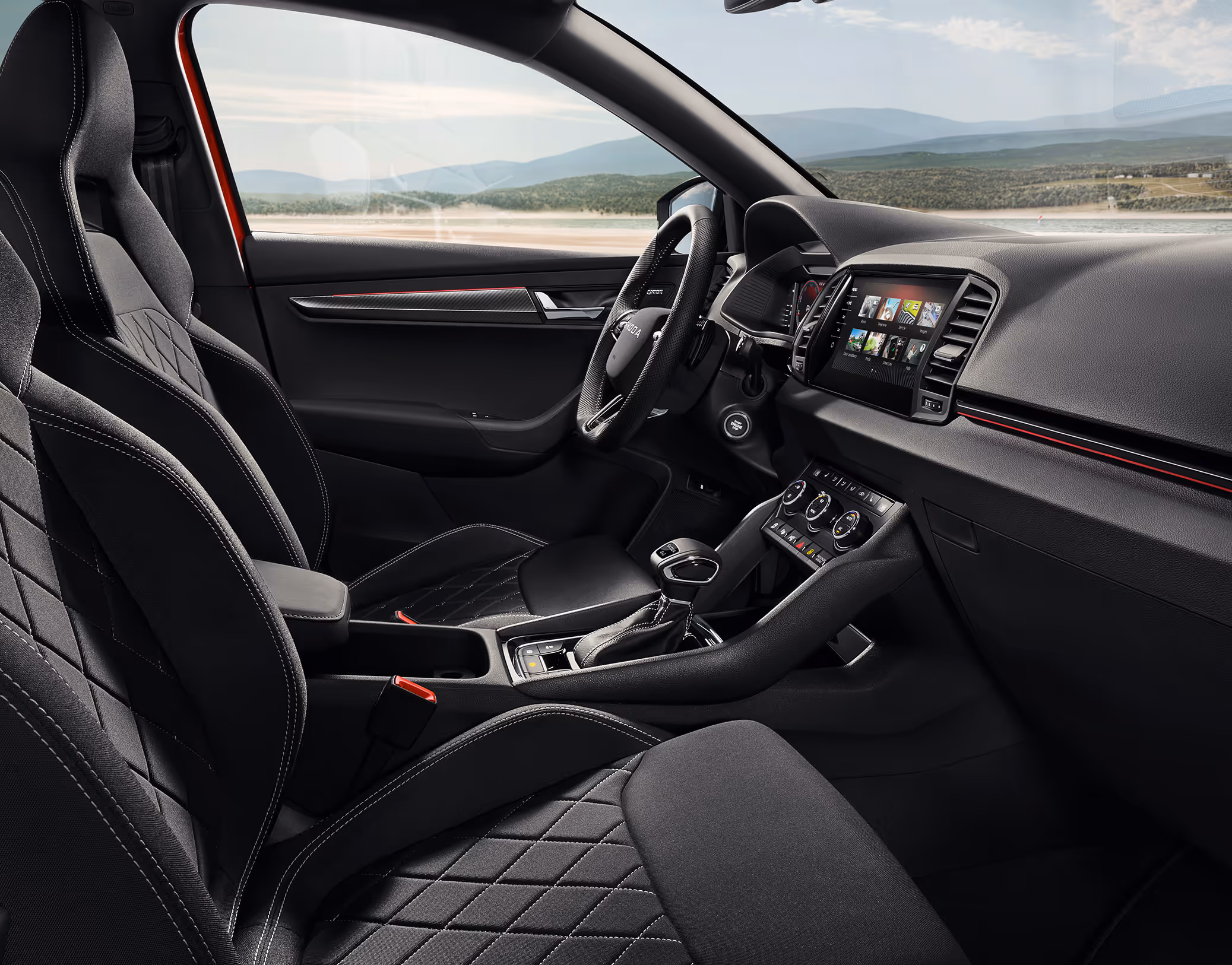 Eine seitliche Innenansicht des Škoda Karoq zeigt das Interieur der Design Selection Sportline. Die schwarzen Sportsitze aus ThermoFlux-Stoff mit integrierten Kopfstützen sowie das dunkle Armaturenbrett mit einer Dekorleiste in Carbon-Optik sind zu sehen, während durch die Fenster eine hügelige, bewaldete Landschaft sichtbar ist.