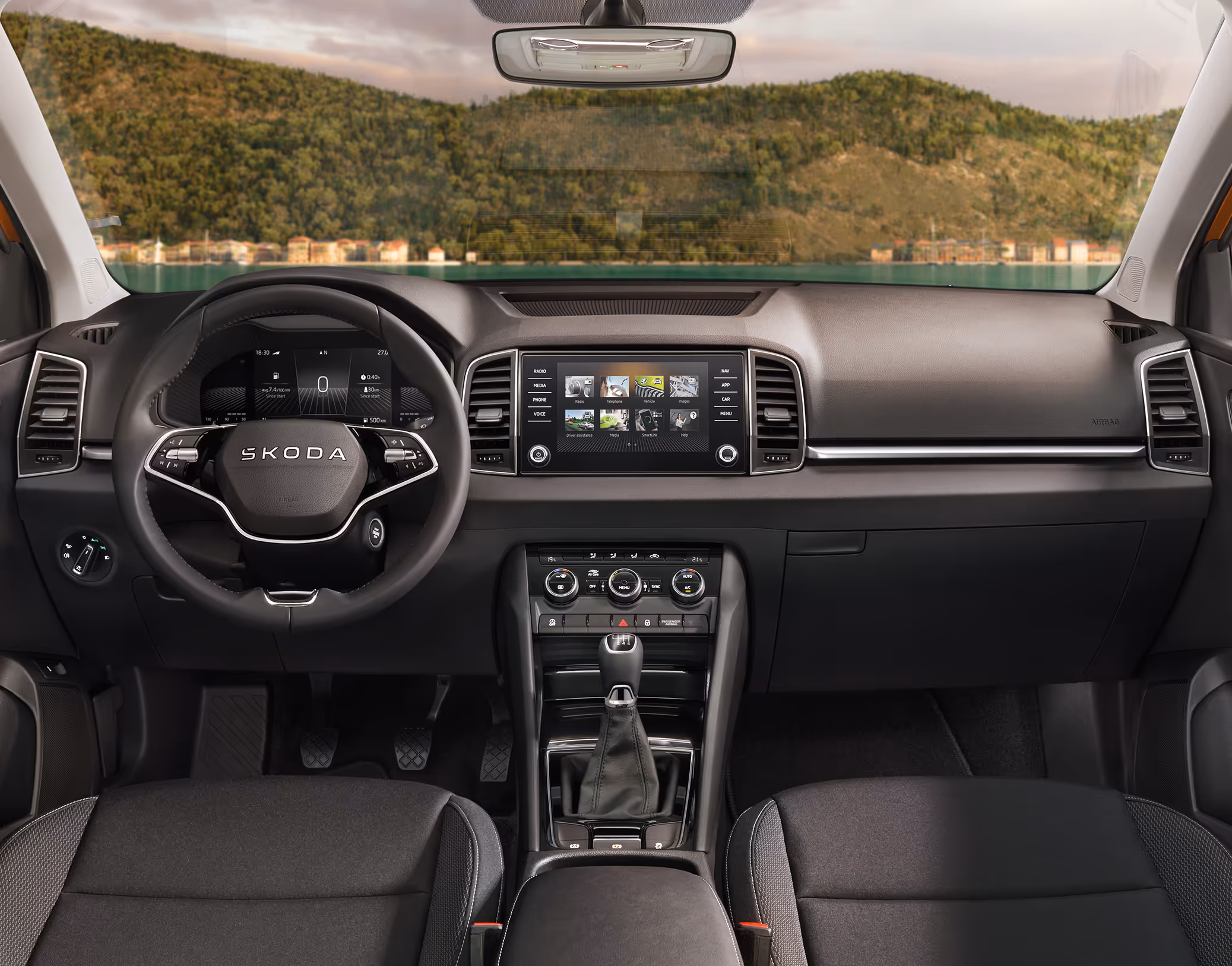 Eine weite Innenansicht zeigt das gesamte Cockpit eines Škoda Karoq mit der Design Selection Loft. Das dunkle Interieur ist mit Stoffsitzen mit grauen Akzenten und einer hellgrauen, strukturierten Dekorleiste am Armaturenbrett ausgestattet. Durch die Windschutzscheibe ist eine Küstenlandschaft mit Hügeln und einem Gewässer zu sehen.