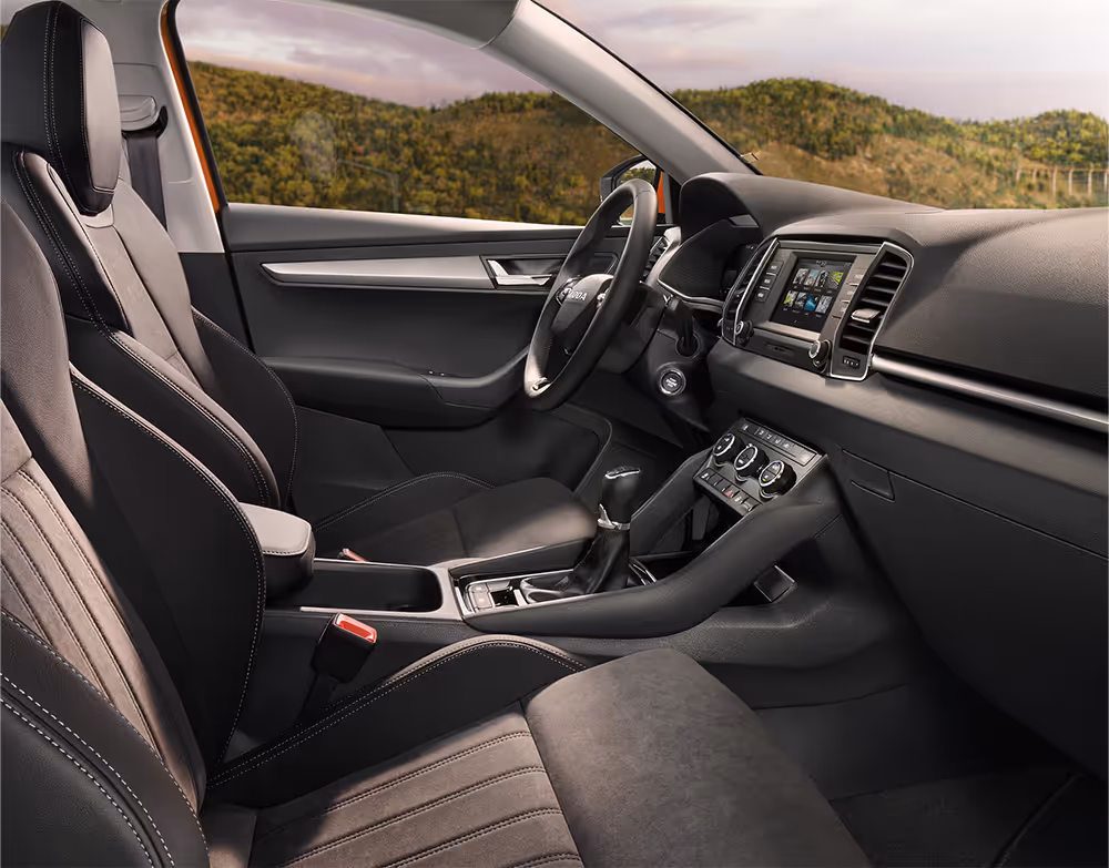 Eine seitliche Innenansicht des Škoda Karoq zeigt das Interieur der Design Selection Lounge. Die schwarzen Suedia-Sitzbezüge mit grauen Kontrastnähten sowie das dunkle Armaturenbrett mit einer Dekorleiste in Piano-Schwarz sind zu sehen, während durch die Fenster eine hügelige, bewaldete Landschaft sichtbar ist.