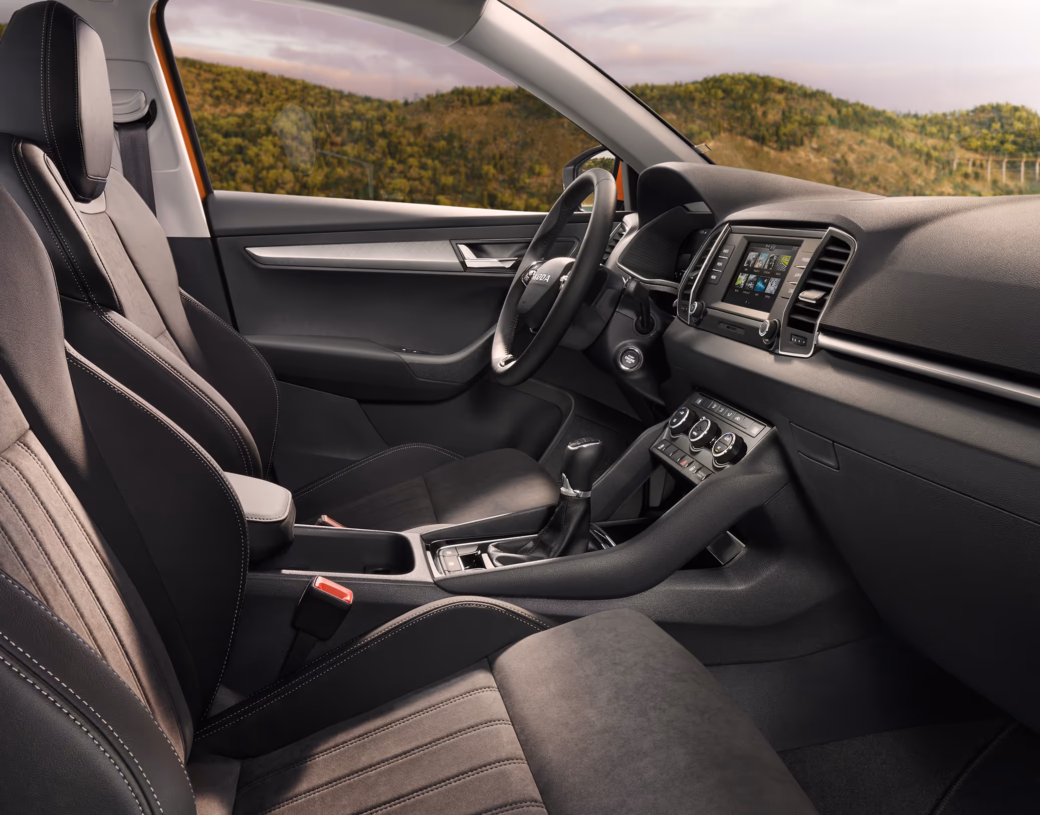 Eine seitliche Innenansicht des Škoda Karoq zeigt das Interieur der Design Selection Lounge. Die schwarzen Suedia-Sitzbezüge mit grauen Kontrastnähten sowie das dunkle Armaturenbrett mit einer Dekorleiste in Piano-Schwarz sind zu sehen, während durch die Fenster eine hügelige, bewaldete Landschaft sichtbar ist.