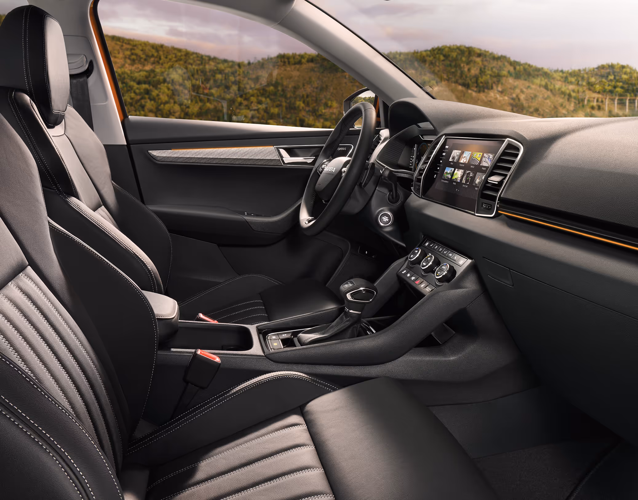 Eine seitliche Innenansicht des Škoda Karoq zeigt das Interieur der Design Selection Suite. Die schwarzen Leder-Kunstleder-Sitzbezüge mit grauen Kontrastnähten sowie das dunkle Armaturenbrett mit einer Dekorleiste in Piano-Schwarz sind zu sehen, während durch die Fenster eine hügelige, bewaldete Landschaft sichtbar ist.