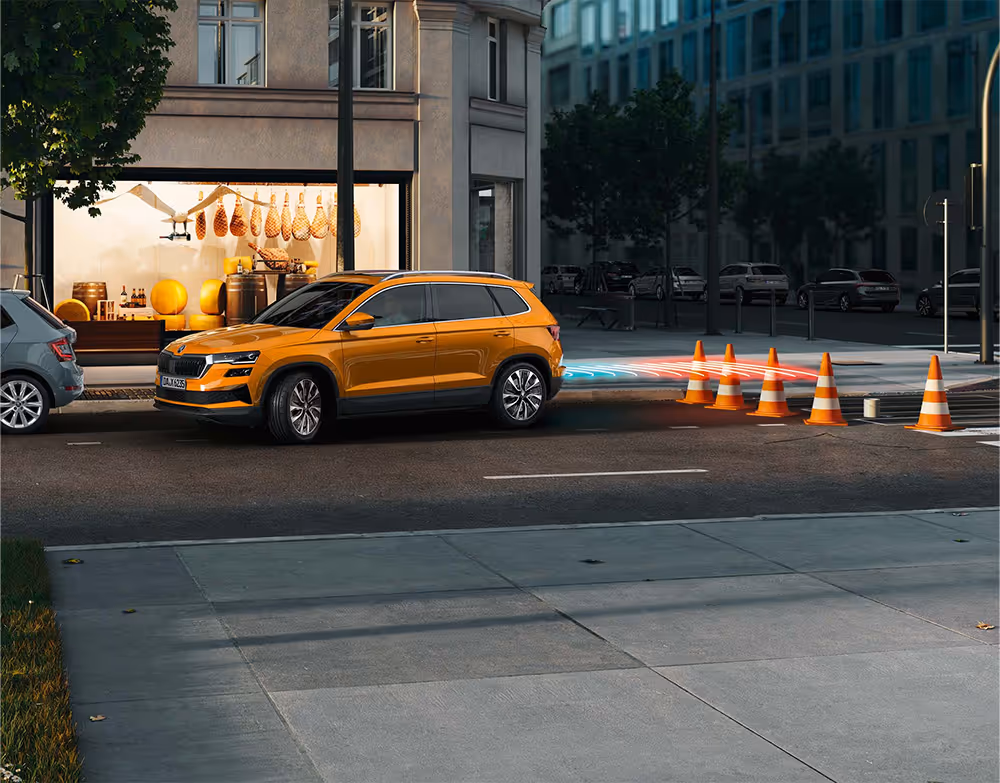 Animation des Rangierbremsassistenten: Ein orangefarbener Škoda Karoq manövriert rückwärts auf eine Reihe von Pylonen zu und bremst automatisch ab, um eine Kollision zu verhindern. Leuchtende rote und blaue Linien visualisieren den vom Sensor erfassten Bereich und die von dem Hindernis ausgehende Gefahr.