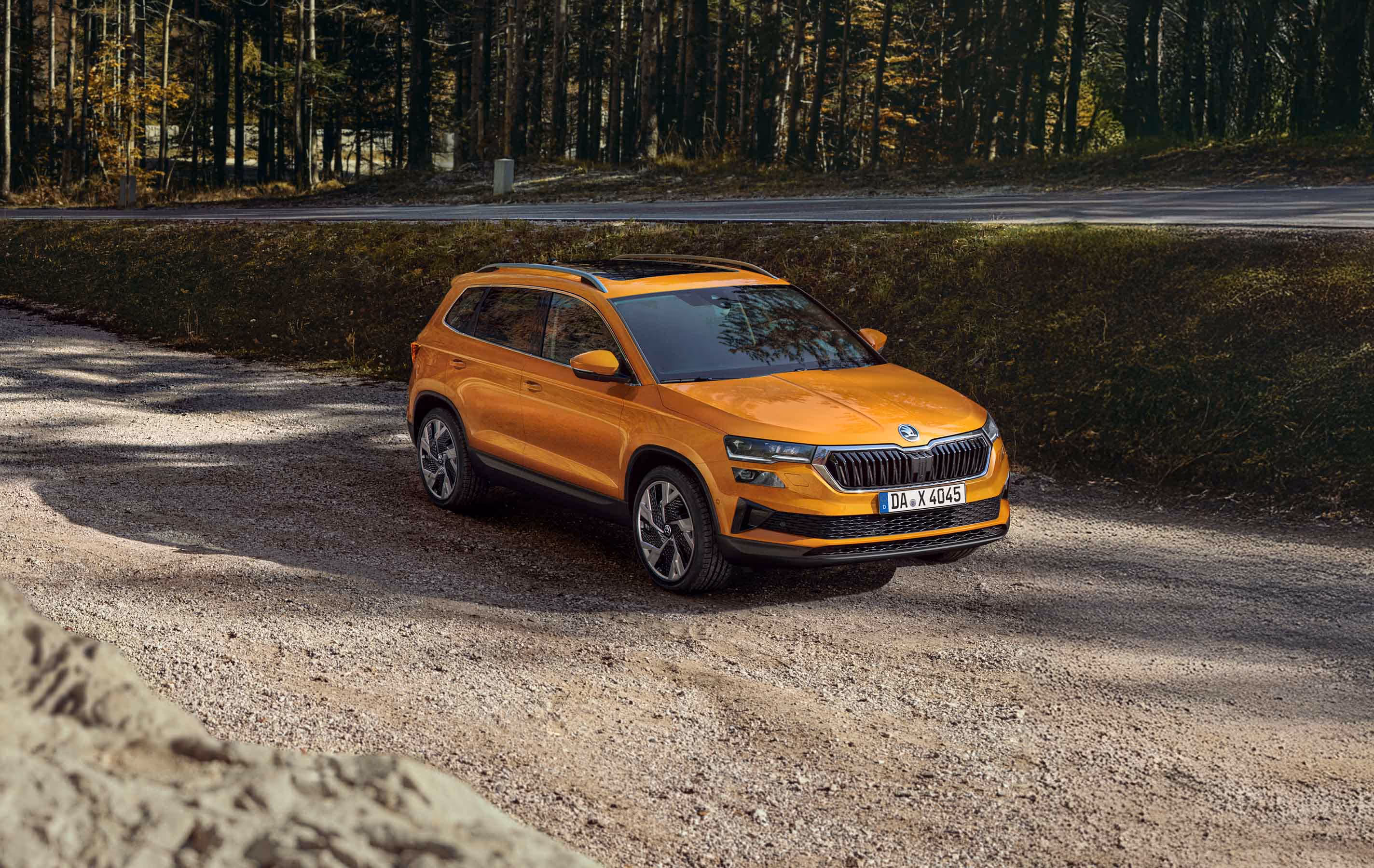 Ein Škoda Karoq in der Lackierung Phoenix-Orange Metallic ist auf einem Schotterplatz vor einem dichten Wald geparkt. Die Aufnahme aus einer leicht erhöhten Dreiviertelperspektive von vorne zeigt das Fahrzeug mit Panoramaschiebedach und silberner Dachreling. Im Hintergrund ist eine asphaltierte Straße zu sehen, die am Waldrand entlangführt.