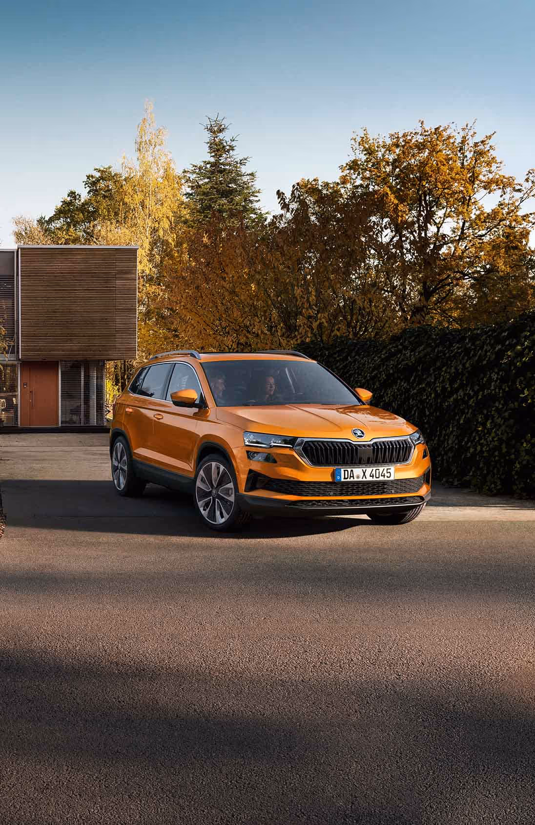 Ein Škoda Karoq in der Lackierung Phoenix-Orange Premium Metallic fährt aus der Einfahrt eines modernen Wohnhauses. Das Fahrzeug wird in einer frontalen Dreiviertelansicht gezeigt, umgeben von Bäumen mit herbstlichem Laub und einer großen, dunklen Hecke. Unten rechts steht auf einer dunkelgrün abgesetzten Fläche "Karoq" geschrieben.