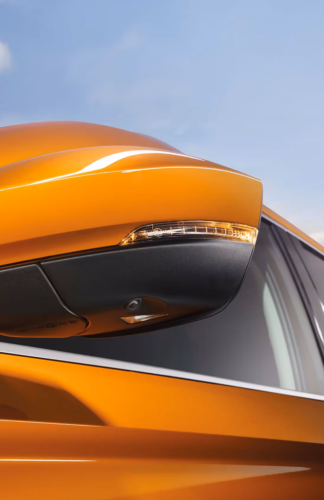 Eine Nahaufnahme zeigt den Außenspiegel eines Škoda Karoq in der Lackierung Phoenix-Orange Metallic. Das Spiegelgehäuse ist in Wagenfarbe lackiert und verfügt über einen integrierten Blinker. Unterhalb des Spiegels sind die Kamera für das Area-View-System und die Einstiegsbeleuchtung zu erkennen. 