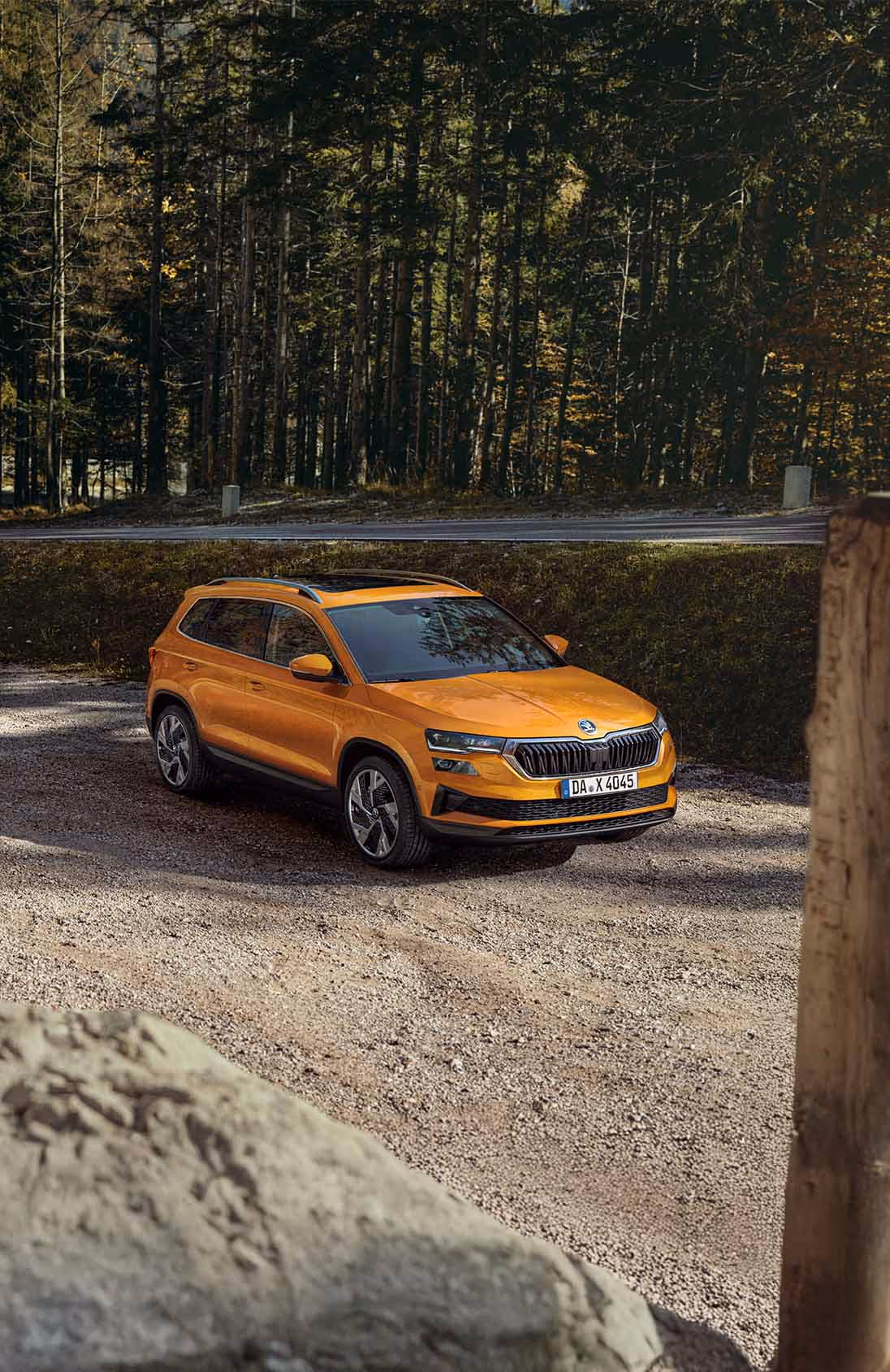 Ein Škoda Karoq in der Lackierung Phoenix-Orange Metallic ist auf einem Schotterplatz vor einem dichten Wald geparkt. Die Aufnahme aus einer leicht erhöhten Dreiviertelperspektive von vorne zeigt das Fahrzeug mit Panoramaschiebedach und silberner Dachreling. Im Hintergrund ist eine asphaltierte Straße zu sehen, die am Waldrand entlangführt.