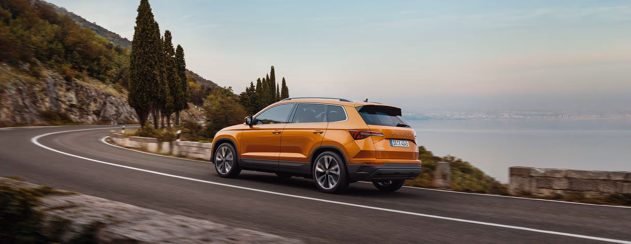 Ein Škoda Karoq in der Lackierung Phoenix-Orange Metallic fährt auf einer kurvenreichen Küstenstraße. Das Fahrzeug wird von schräg hinten gezeigt, während es an Zypressen und einer hügeligen Landschaft mit Blick auf das Meer vorbeifährt.