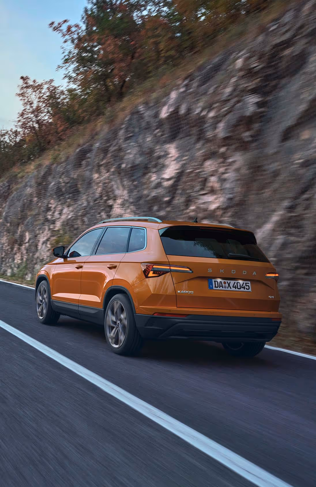 Ein Škoda Karoq in der Lackierung Phoenix-Orange Metallic fährt auf einer asphaltierten Straße. Das Fahrzeug wird von schräg hinten gezeigt, während es an einer felsigen, bewachsenen Steilwand entlangfährt. Die Bewegungsunschärfe des Hintergrunds und der Räder unterstreicht die Fahrdynamik.