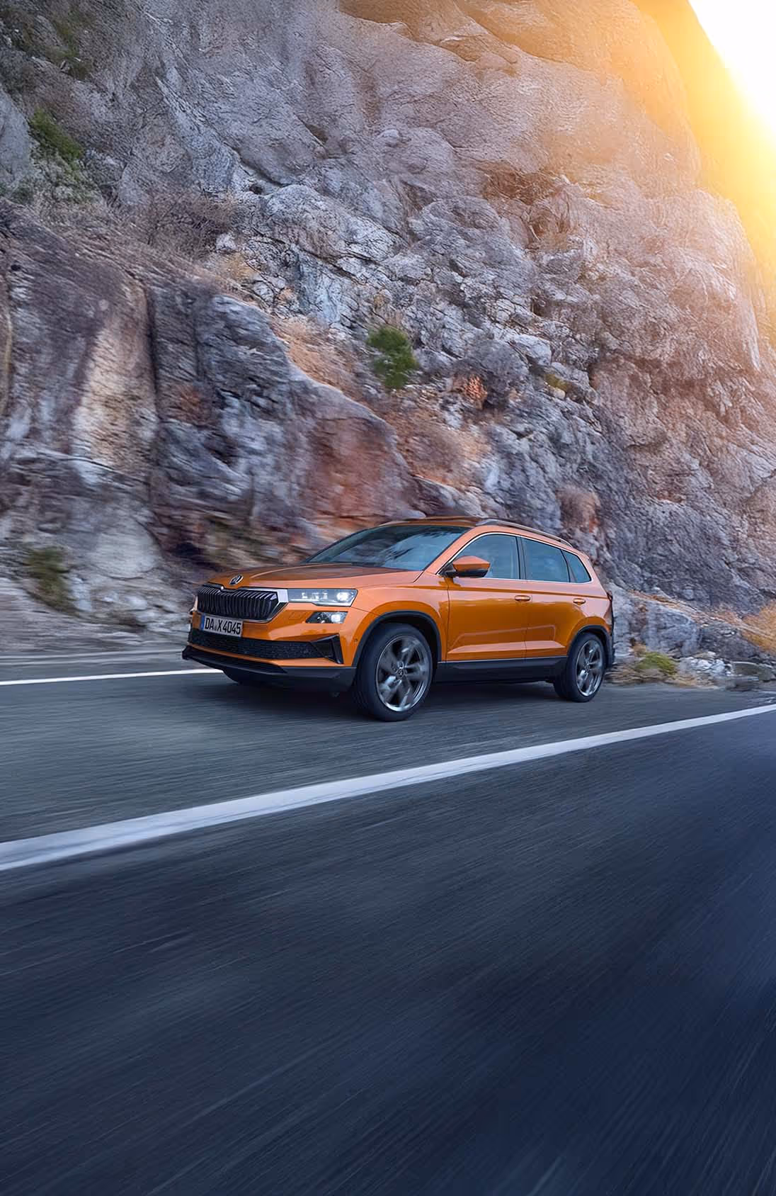 Ein Škoda Karoq in der Lackierung Phoenix-Orange Metallic fährt auf einer kurvenreichen Straße entlang einer Felswand. Das Fahrzeug wird in einer dynamischen Dreiviertelansicht von vorne gezeigt. Die Bewegungsunschärfe des felsigen Hintergrunds verdeutlicht die Fahrszene. Unten rechts steht auf einer dunkelgrün abgesetzten Fläche "Škoda Connect" geschrieben.