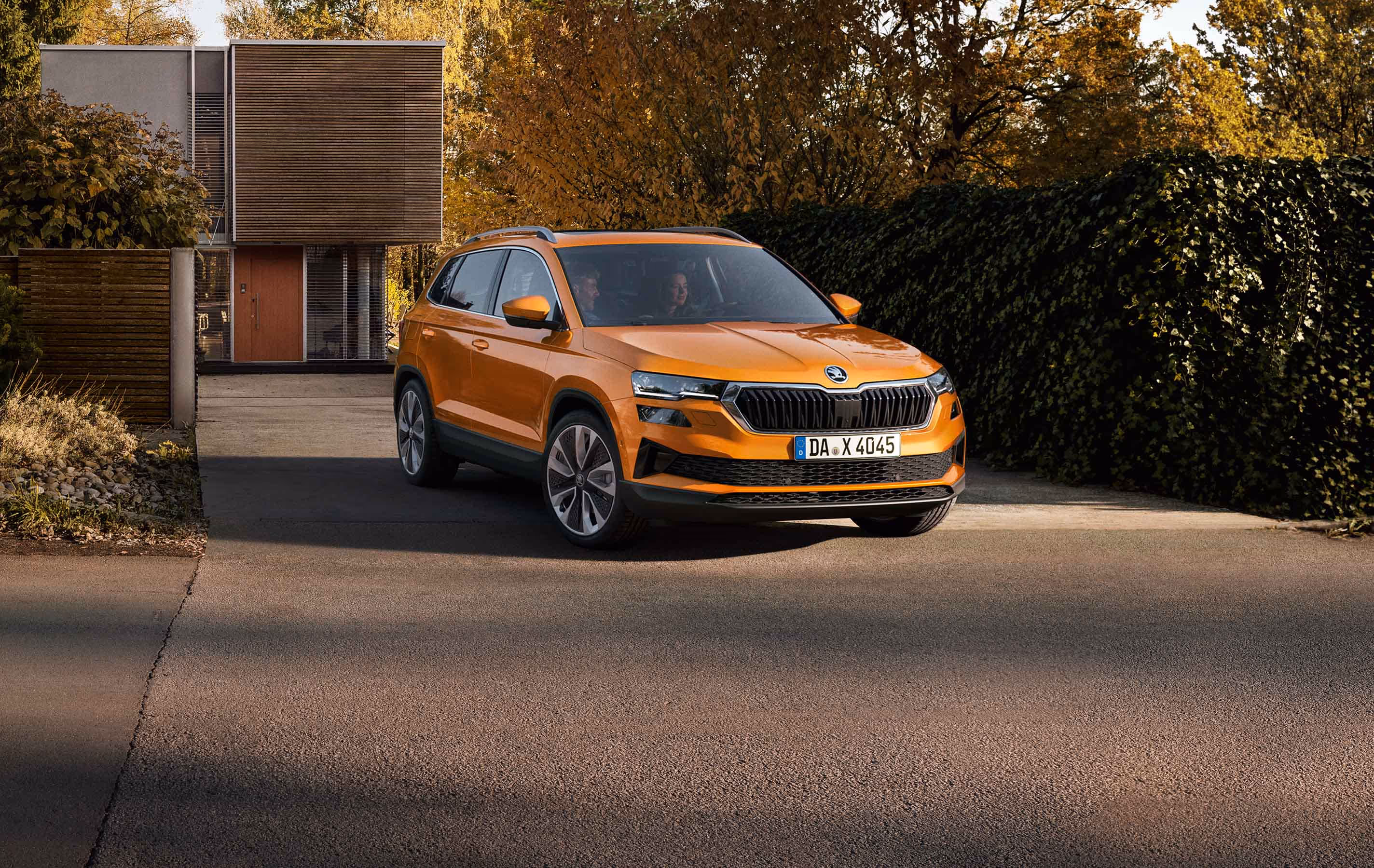 Ein Škoda Karoq in der Lackierung Phoenix-Orange Metallic fährt aus der Einfahrt eines modernen Wohnhauses. Das Fahrzeug wird in einer frontalen Dreiviertelansicht gezeigt, umgeben von Bäumen mit herbstlichem Laub und einer großen, dunklen Hecke. 