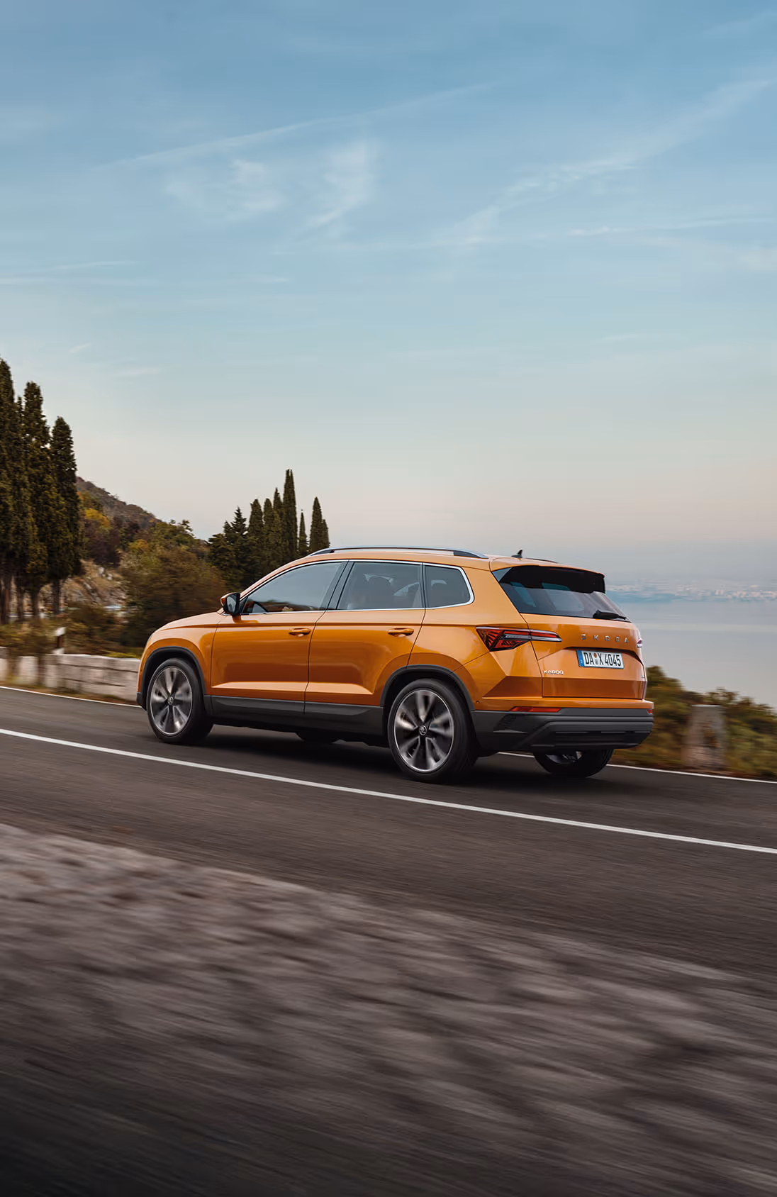 Ein Škoda Karoq in der Lackierung Phoenix-Orange Metallic fährt auf einer kurvenreichen Küstenstraße. Das Fahrzeug wird von schräg hinten gezeigt, während es an Zypressen und einer hügeligen Landschaft mit Blick auf das Meer vorbeifährt.