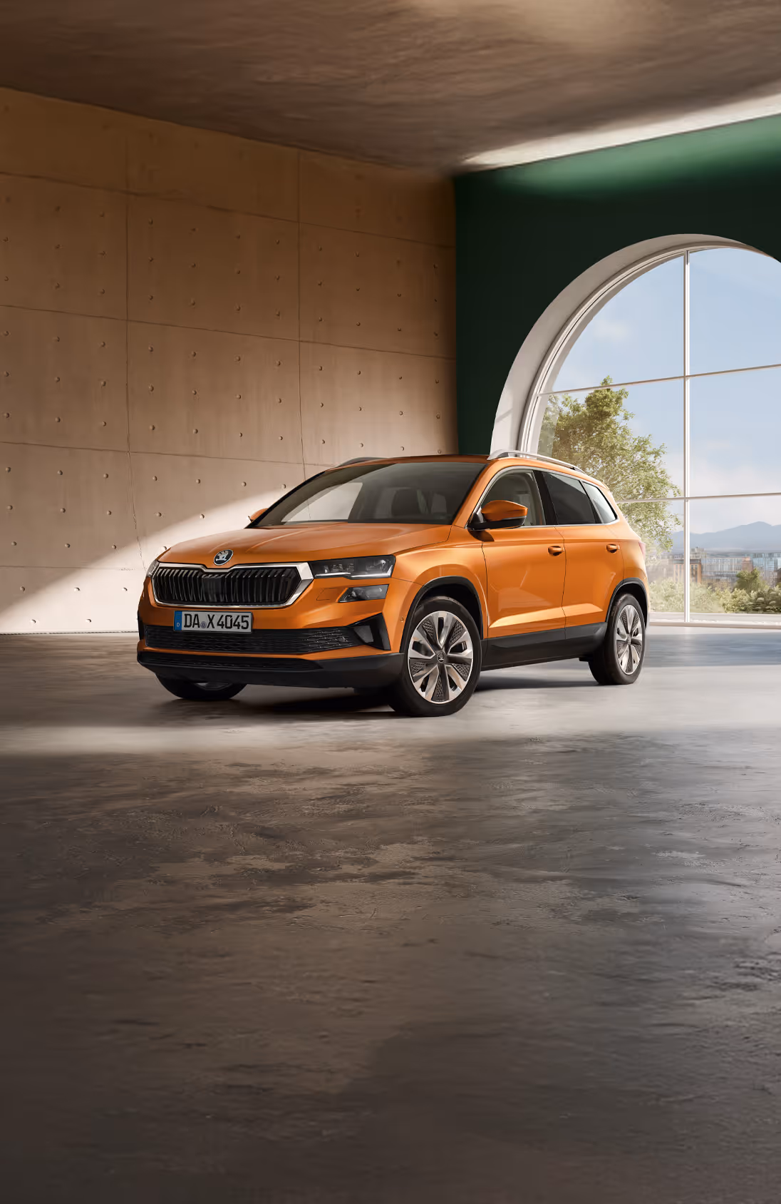 Ein Škoda Karoq in der Lackierung Phoenix-Orange Premium Metallic fährt aus der Einfahrt eines modernen Wohnhauses. Das Fahrzeug wird in einer frontalen Dreiviertelansicht gezeigt, umgeben von Bäumen mit herbstlichem Laub und einer großen, dunklen Hecke. Unten rechts steht auf einer dunkelgrün abgesetzten Fläche "Karoq" geschrieben.