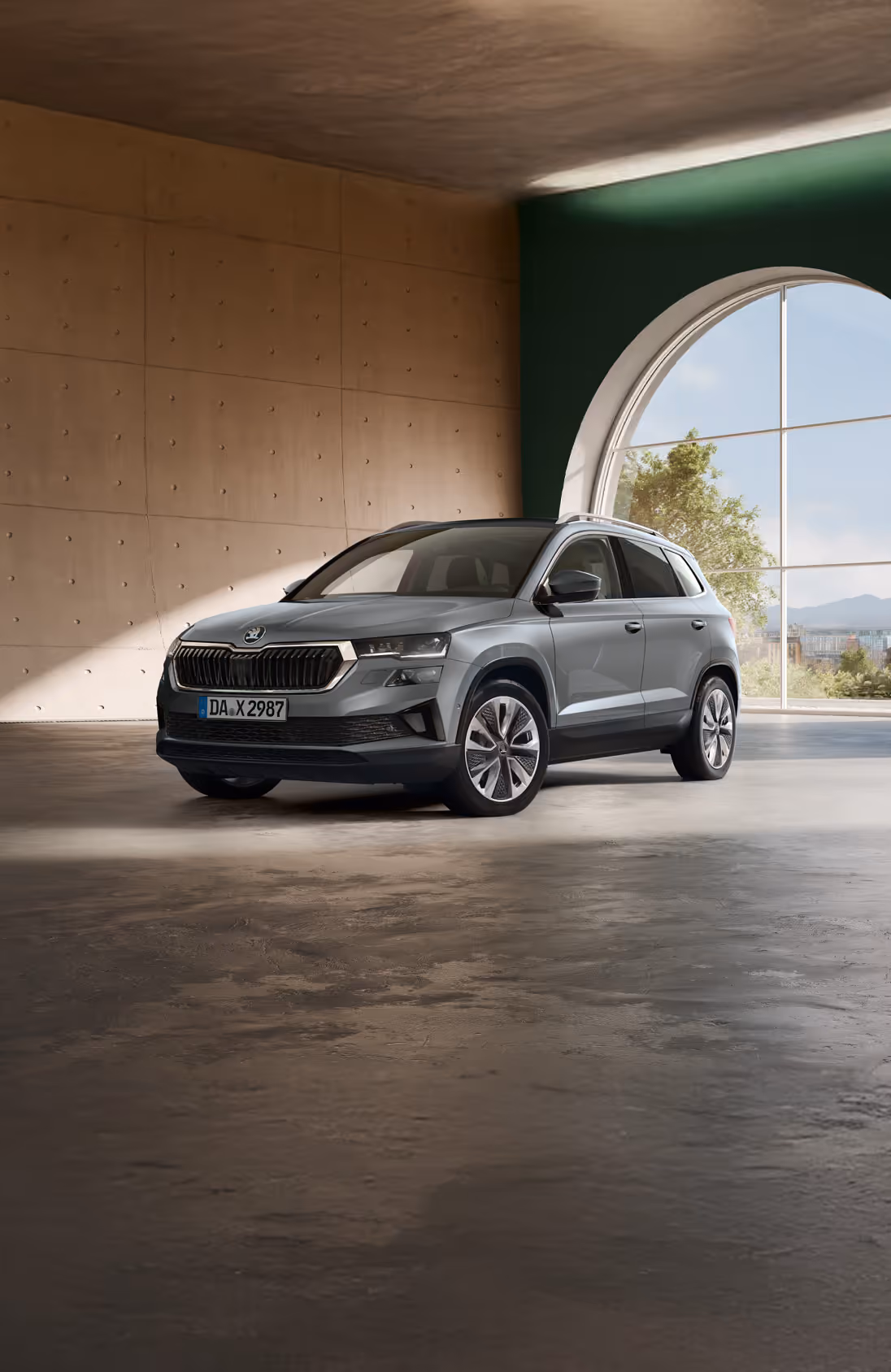 Ein Škoda Karoq in der Lackierung Phoenix-Orange Premium Metallic fährt aus der Einfahrt eines modernen Wohnhauses. Das Fahrzeug wird in einer frontalen Dreiviertelansicht gezeigt, umgeben von Bäumen mit herbstlichem Laub und einer großen, dunklen Hecke. Unten rechts steht auf einer dunkelgrün abgesetzten Fläche "Karoq" geschrieben.