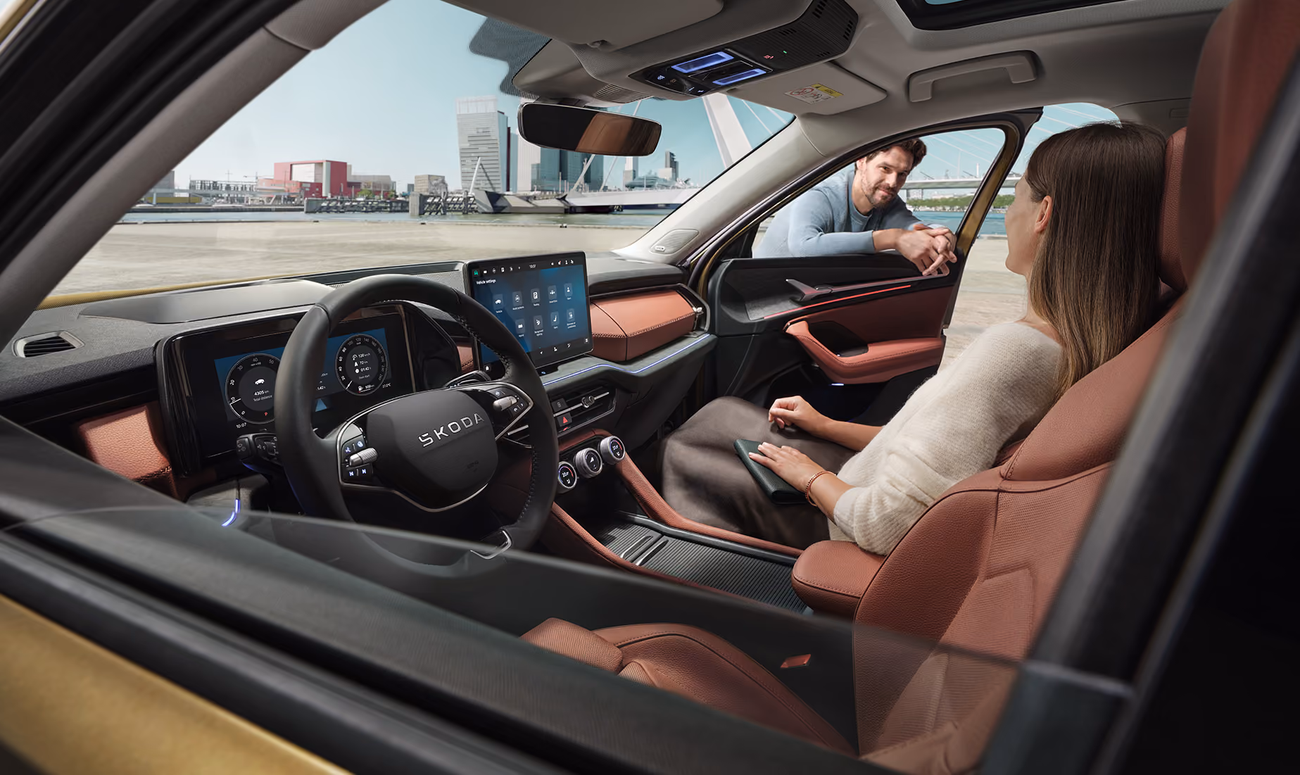 Ein Blick aus dem Inneren eines Škoda Kodiaq zeigt eine Frau auf dem Beifahrersitz, die sich mit einem Mann unterhält, der sich durch das offene Fenster lehnt. Die Szene hebt das Interieur der Design Selection Lounge mit cognac-braunen Elementen, das große, freistehende 13-Zoll-Infotainmentdisplay und das digitale Cockpit hervor. Durch die Windschutzscheibe ist eine moderne Stadtkulisse an einem Kai zu sehen, was eine helle und offene Atmosphäre schafft. Unten recht im Bild steht auf einer dunkelgrün abgesetzten Fläche "Smarte Details" geschrieben.