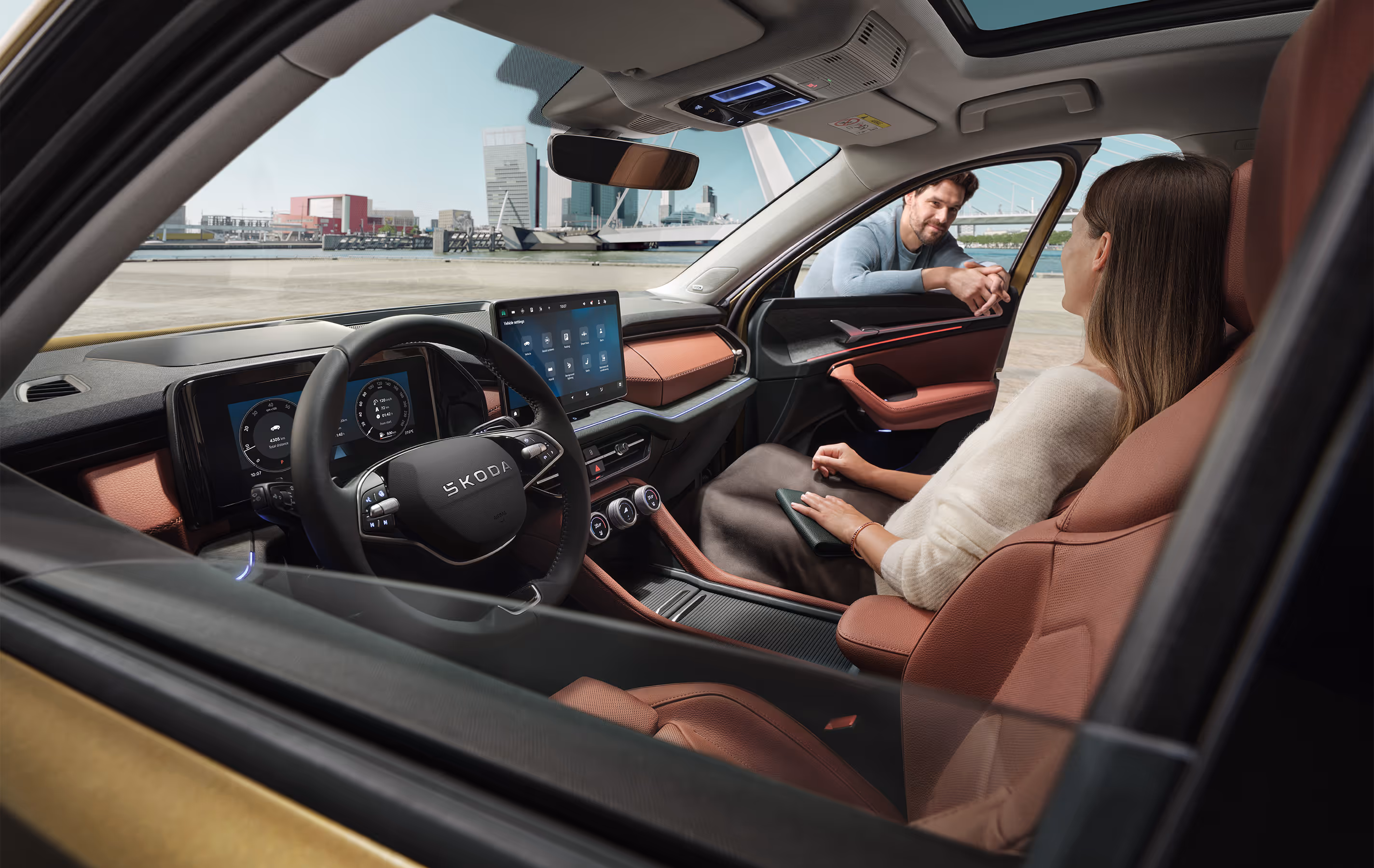Ein Blick aus dem Inneren eines Škoda Kodiaq zeigt eine Frau auf dem Beifahrersitz, die sich mit einem Mann unterhält, der sich durch das offene Fenster lehnt. Die Szene hebt das Interieur der Design Selection Lounge mit cognac-braunen Elementen, das große, freistehende 13-Zoll-Infotainmentdisplay und das digitale Cockpit hervor. Durch die Windschutzscheibe ist eine moderne Stadtkulisse an einem Kai zu sehen, was eine helle und offene Atmosphäre schafft. Unten recht im Bild steht auf einer dunkelgrün abgesetzten Fläche "Smarte Details" geschrieben.