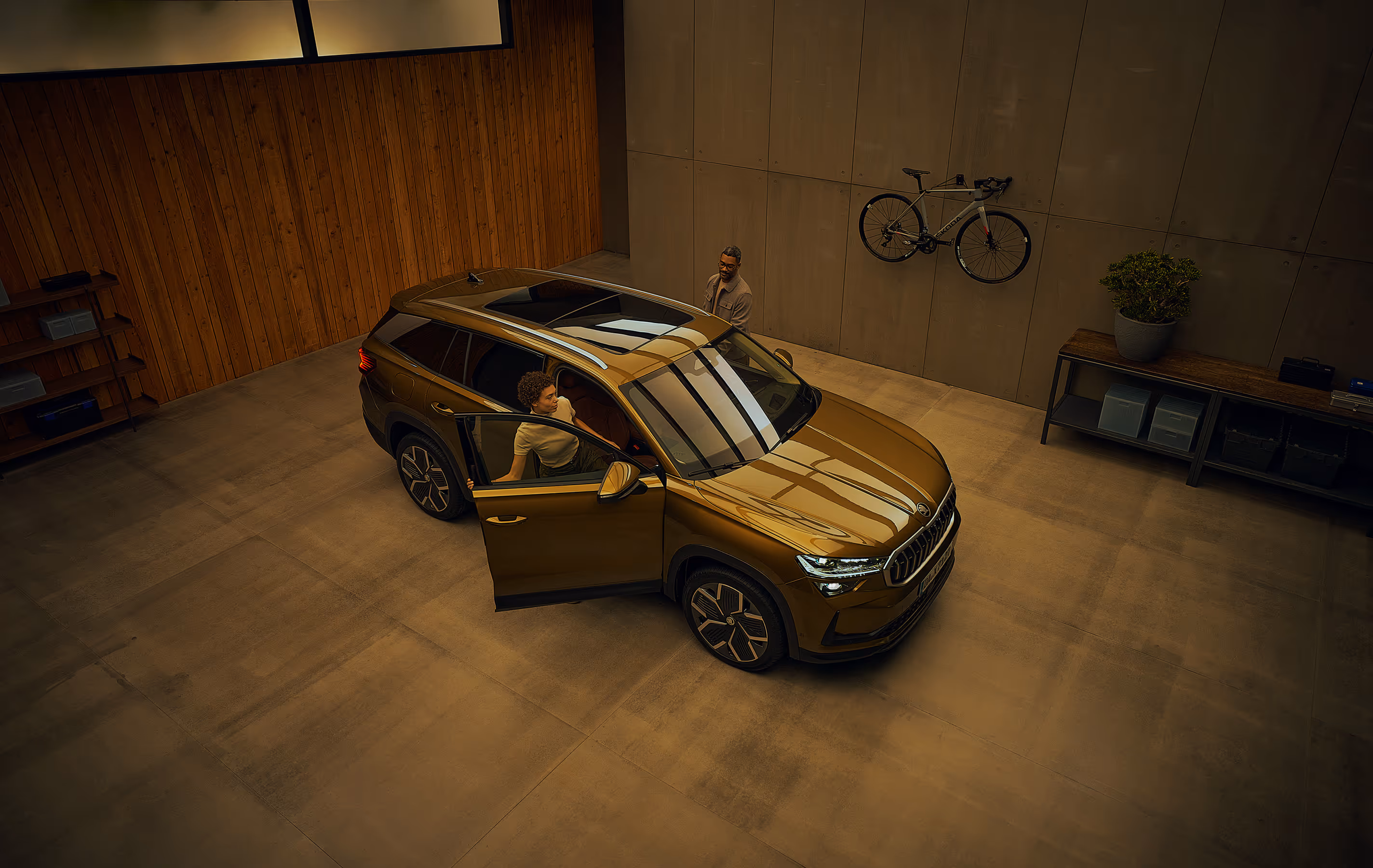 Blick aus einer erhöhten Perspektive auf einen Škoda Kodiaq in der Lackierung Gold-Bronze Metallic, der in einer modernen Garage parkt. Eine Frau steigt auf der Beifahrerseite ein, während ein Mann gleichzeitig die Fahrertür öffnet. An der Garagenwand ist ein sportliches Škoda Fahrrad mit silbernem Rahmen befestigt. Die Deckenbeleuchtung spiegelt sich auf dem Fahrzeugdach, der Windschutzscheibe und der Motorhaube.