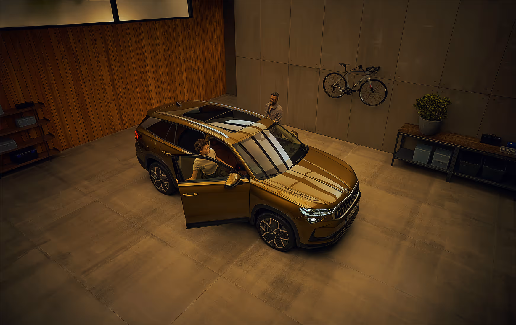 Blick aus einer erhöhten Perspektive auf einen Škoda Kodiaq in der Lackierung Gold-Bronze Metallic, der in einer modernen Garage parkt. Eine Frau steigt auf der Beifahrerseite ein, während ein Mann gleichzeitig die Fahrertür öffnet. An der Garagenwand ist ein sportliches Škoda Fahrrad mit silbernem Rahmen befestigt. Die Deckenbeleuchtung spiegelt sich auf dem Fahrzeugdach, der Windschutzscheibe und der Motorhaube.