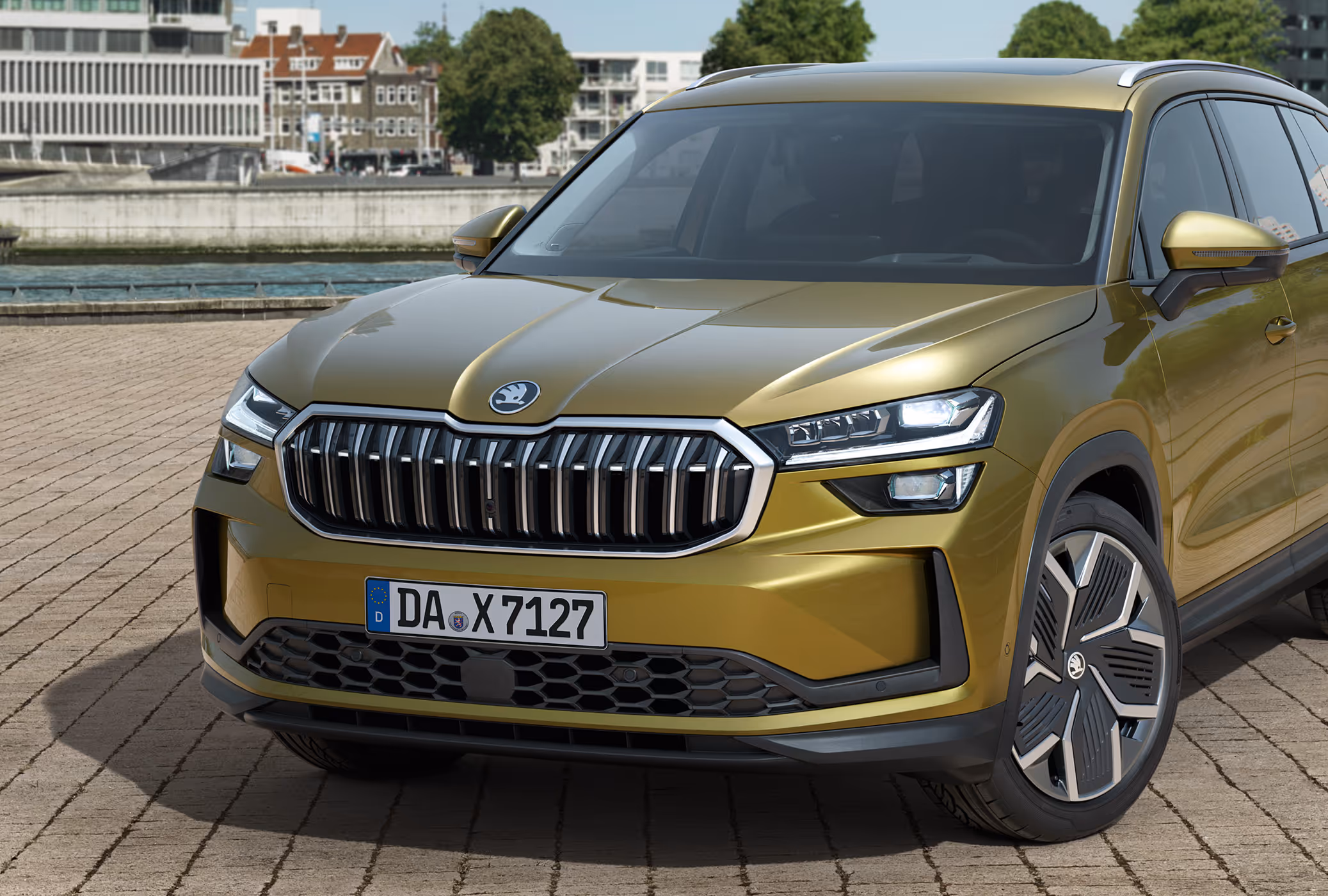Eine Nahaufnahme zeigt die Frontpartie eines Škoda Kodiaq in Gold-Bronze Metallic, der auf einem gepflasterten Platz an einem Kai steht. Die Perspektive hebt das Design der Fahrzeugfront hervor, insbesondere den markanten, hexagonalen Škoda Kühlergrill und die LED-Matrix-Scheinwerfer. Im unscharfen Hintergrund sind Gebäude und Bäume am gegenüberliegenden Ufer zu sehen.
