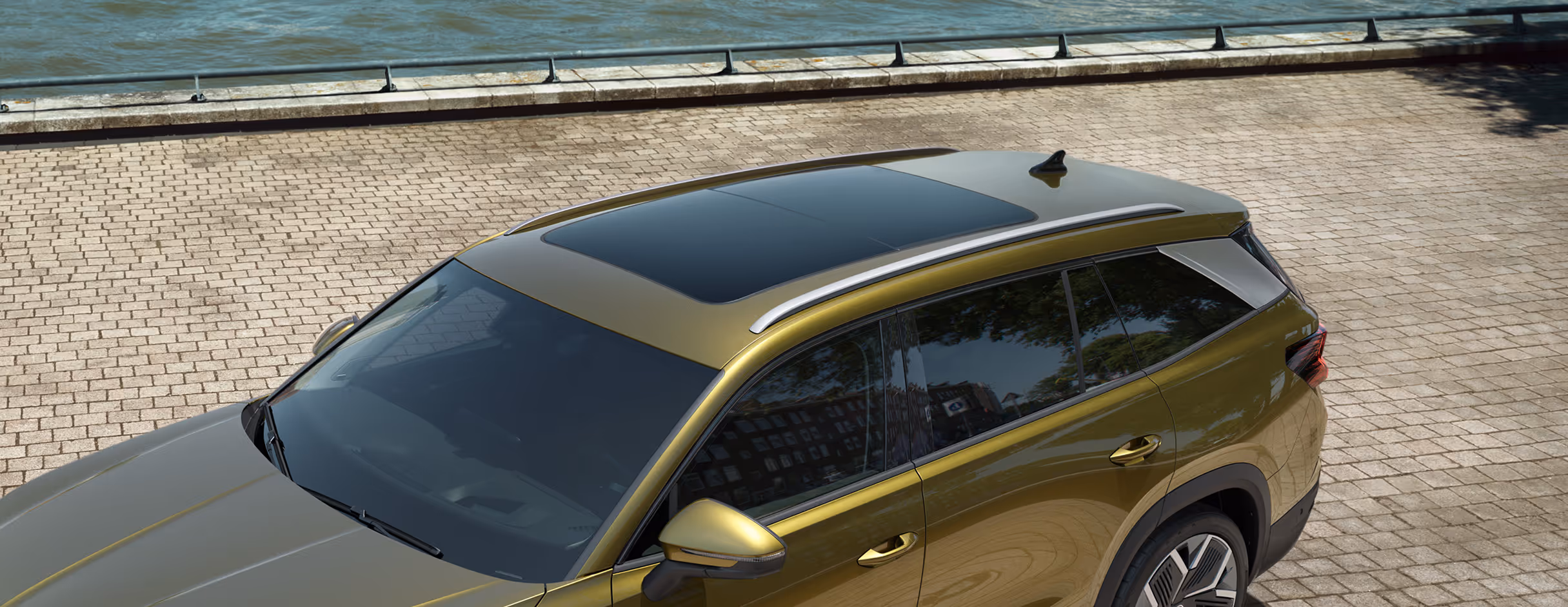 Ein Škoda Kodiaq in der Lackierung Gold-Bronze Metallic wird aus einer erhöhten Perspektive gezeigt, die den Blick auf das Dach lenkt. Im Fokus stehen das große, optionale Panoramaschiebedach, die silberne Dachreling und das Zierelement an der D-Säule. Das Fahrzeug parkt auf einem gepflasterten Kai neben einem Gewässer.