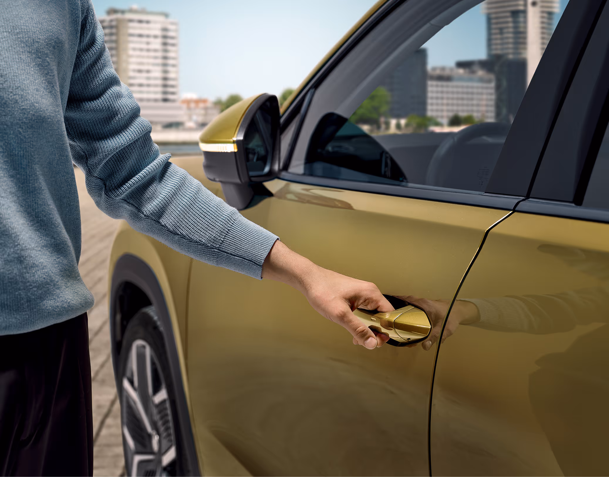 Eine Nahaufnahme zeigt, wie eine Person die Tür eines Škoda Kodiaq in Gold-Bronze Metallic öffnet. Die Hand greift den Türgriff, um das schlüssellose Zugangs- und Start-Stopp-System KESSY zu nutzen. Die Szene spielt sich in einer städtischen Umgebung ab, die im unscharfen Hintergrund zu erkennen ist.