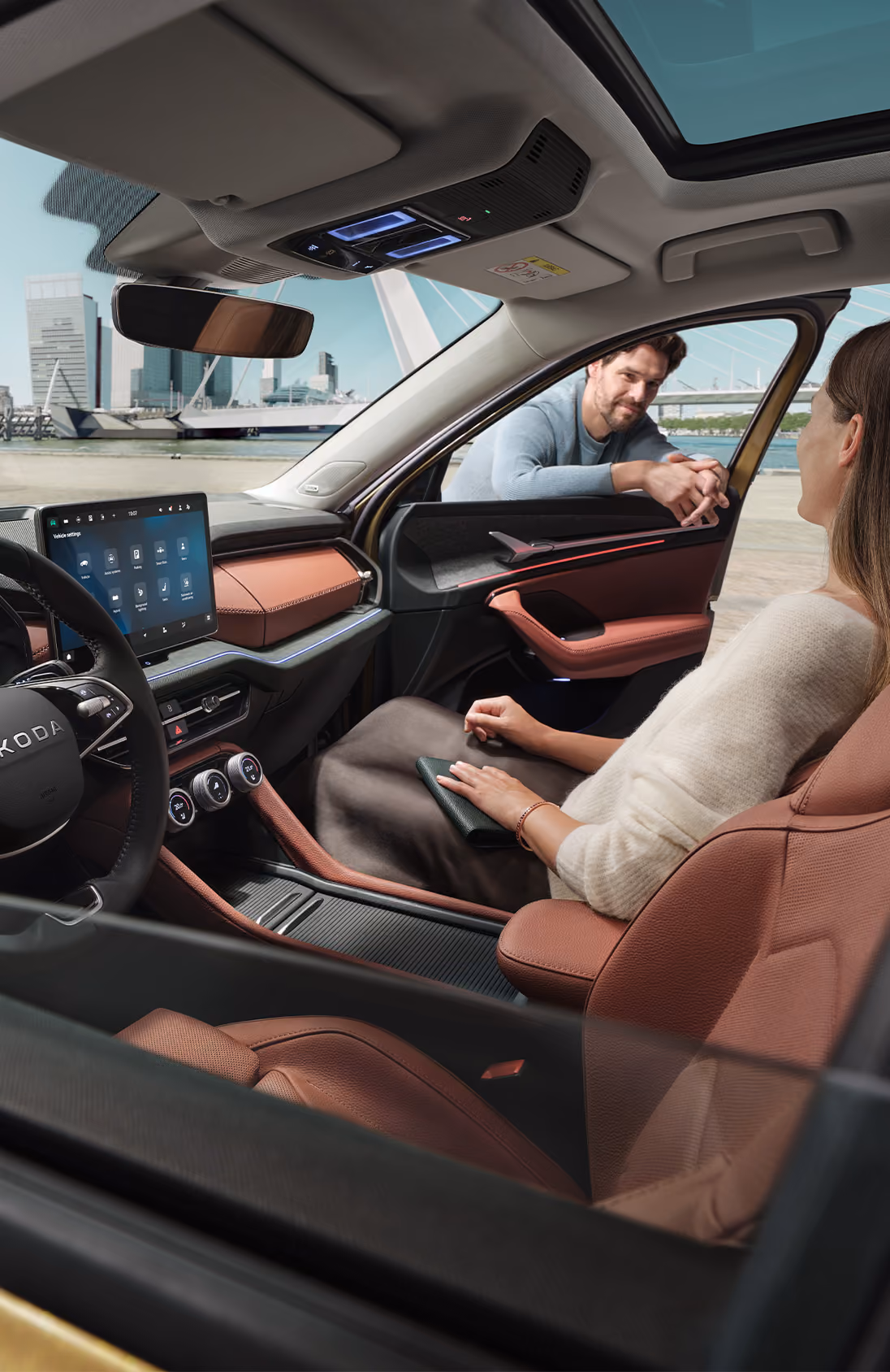 Ein Blick aus dem Inneren eines Škoda Kodiaq zeigt eine Frau auf dem Beifahrersitz, die sich mit einem Mann unterhält, der sich durch das offene Fenster lehnt. Die Szene hebt das Interieur der Design Selection Lounge mit cognac-braunen Elementen, das große, freistehende 13-Zoll-Infotainmentdisplay und das digitale Cockpit hervor. Durch die Windschutzscheibe ist eine moderne Stadtkulisse an einem Kai zu sehen, was eine helle und offene Atmosphäre schafft. Unten recht im Bild steht auf einer dunkelgrün abgesetzten Fläche "Smarte Details" geschrieben.