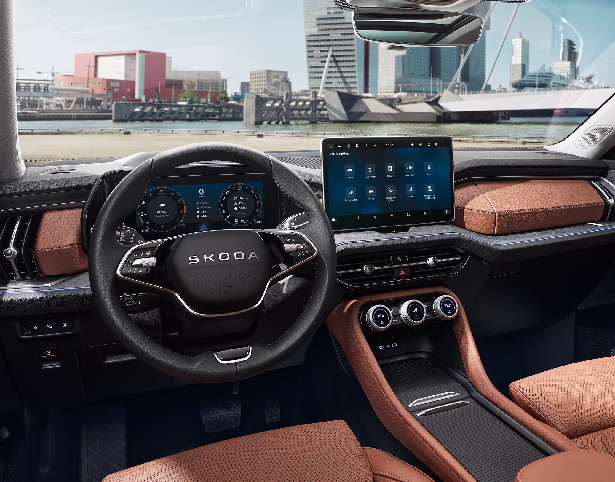 Frontale Ansicht des Cockpits eines Škoda Kodiaq in der Design Selection Lounge in Cognac-Braun. Im Fokus stehen das aufgeräumte Armaturenbrett mit dem freistehenden 13-Zoll-Infotainmentdisplay, dem digitalen Cockpit und den multifunktionalen Smart Dials in der Mittelkonsole. Durch die Windschutzscheibe blickt man auf eine sonnige, moderne Hafenstadt.
