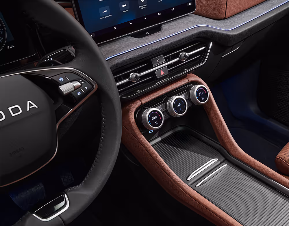 Eine Nahaufnahme der Mittelkonsole im Škoda Kodiaq hebt die drei multifunktionalen Smart Dials mit ihren integrierten Digitalanzeigen hervor. Die Konsole, eingefasst in cognac-braune Elemente der Design Selection Lounge, beherbergt auch die Phonebox mit kabelloser Ladefunktion in einem großen Ablagefach. Oberhalb der Regler sind die Lüftungsdüsen und die blaue Ambientebeleuchtung zu sehen.