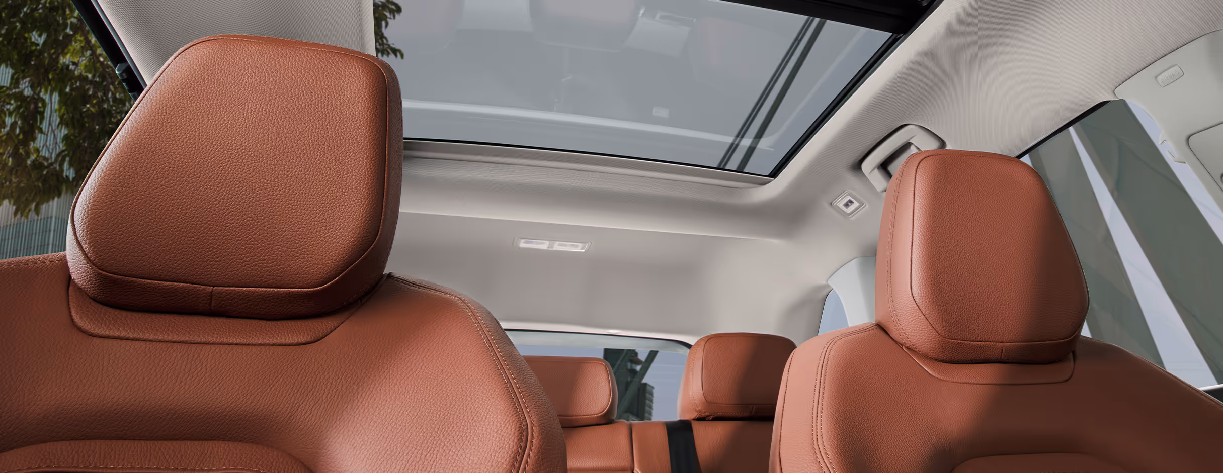 Ein Blick aus dem vorderen Innenraumbereichs eines Škoda Kodiaq nach oben auf das große, optionale Panoramaschiebedach, das den Innenraum mit Tageslicht durchflutet. Die Aufnahme zeigt die Kopfstützen der Vordersitze, die mit cognac-braunem Leder der Design Selection Lounge bezogen sind.