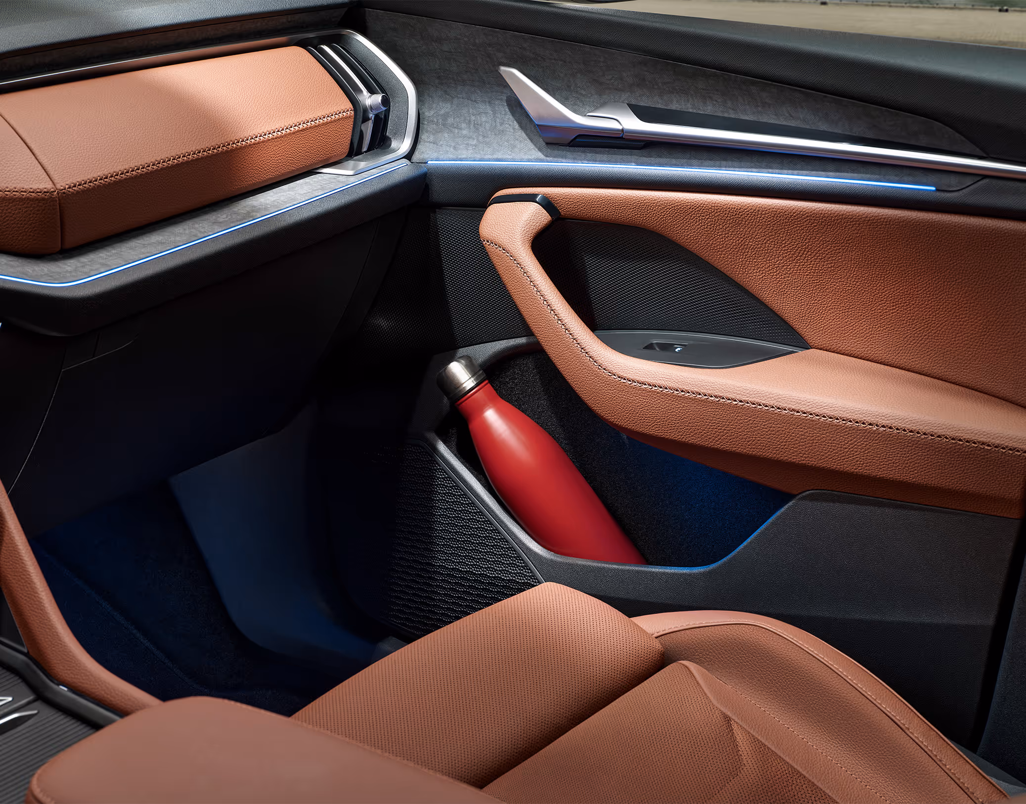 Eine Nahaufnahme zeigt das geräumige Ablagefach in der Türverkleidung eines Škoda Kodiaq, in dem eine rote Trinkflasche Platz findet. Oberhalb des Fachs ist die Armlehne mit dem cognac-braunen Lederbezug der Design Selection Lounge zu sehen.