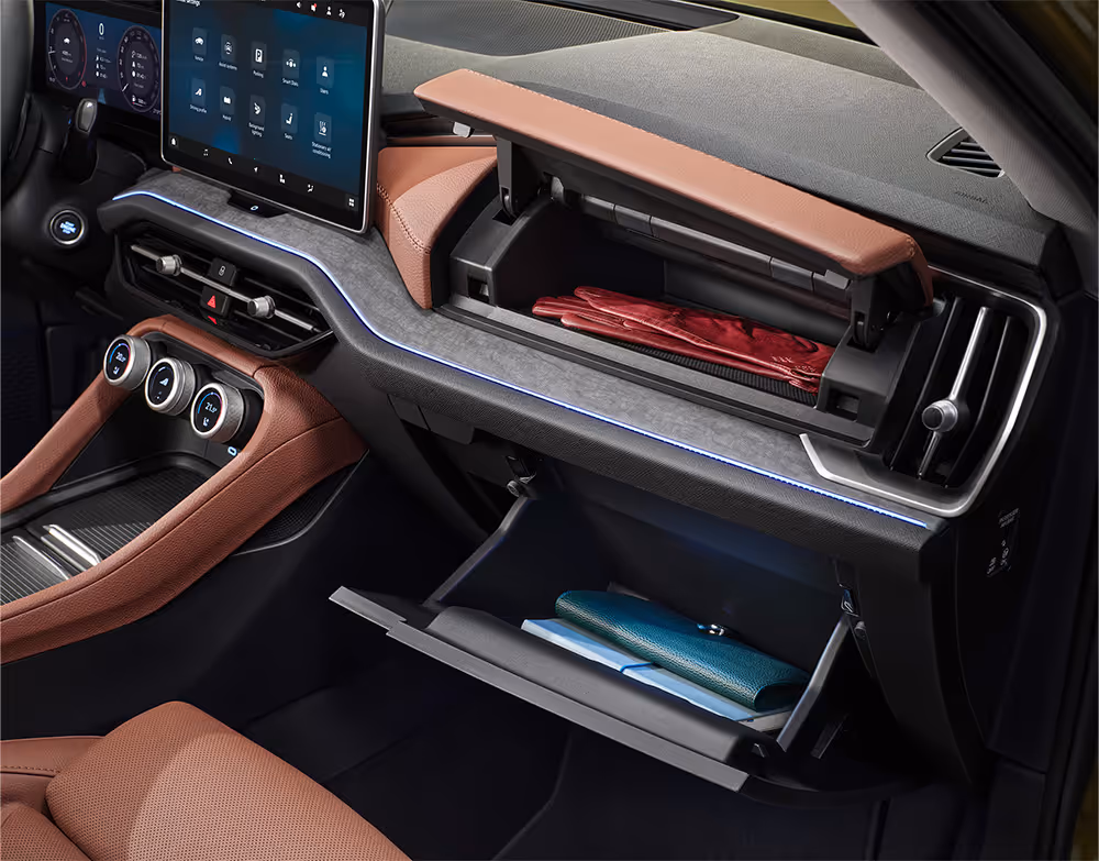 Das Bild zeigt das geöffnete, zweigeteilte Handschuhfach auf der Beifahrerseite eines Škoda Kodiaq. Im oberen Fach liegen rote Handschuhe, während das untere, beleuchtete Fach eine Geldbörse enthält. Die Szene wird durch die blaue Ambientebeleuchtung und die cognac-braunen Akzente der Design Selection Lounge stimmungsvoll in Szene gesetzt.