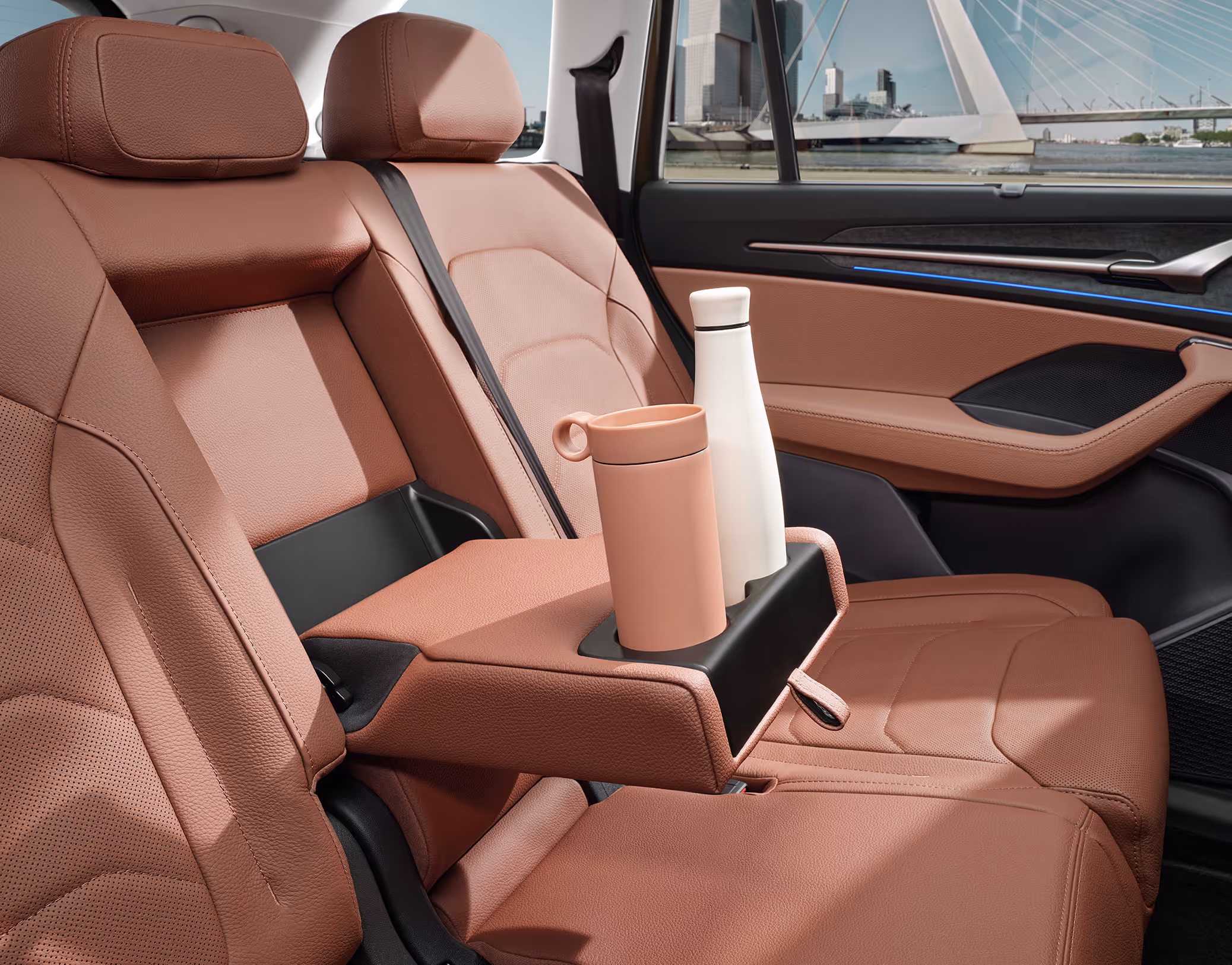 Eine Nahaufnahme zeigt die heruntergeklappte Mittelarmlehne auf de Rückbank eines Škoda Kodiaq. In den integrierten Getränkehaltern stehen ein Thermobecher und eine Trinkflasche. Die Armlehne und die Sitze sind mit dem cognac-braunen Leder der Design Selection Lounge bezogen.