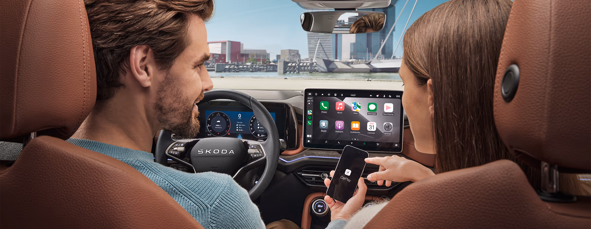 Blick von der Rückbank aus auf ein Paar im vorderen Teil eines Škoda Kodiaq. Der Mann auf dem Fahrersitz hält seiner Partnerin ein Smartphone hin. Die Frau auf dem Beifahrersitz bedient es und die Benutzeroberfläche wird über die kabellose SmartLink-Funktion auf dem großen, zentralen Infotainmentdisplay gespiegelt.
