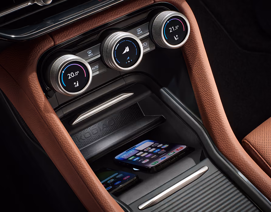 Eine Nahaufnahme zeigt die Phonebox in der Mittelkonsole eines Škoda Kodiaq, in der zwei Smartphones nebeneinander kabellos geladen werden. Das Ablagefach befindet sich direkt unter den Smart Dials und ist von den cognac-braunen Lederelementen der Design Selection Lounge umgeben.