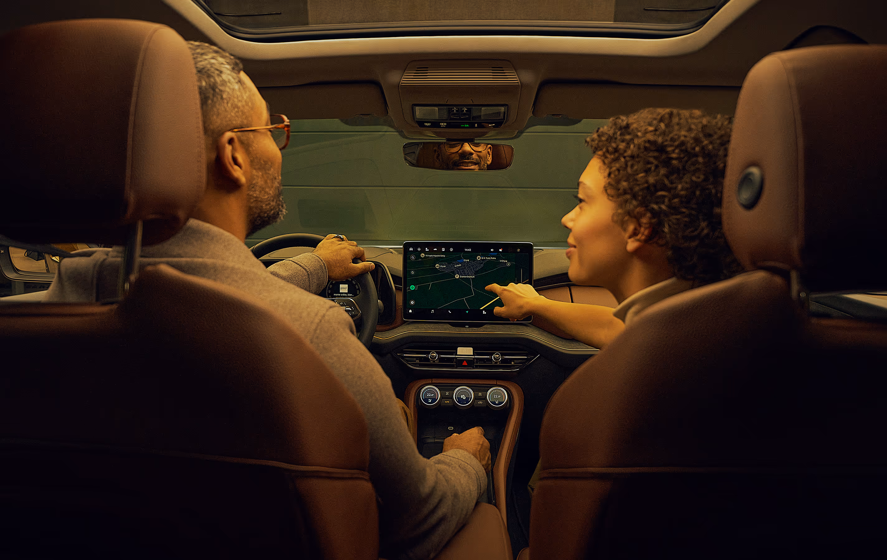 Blick von der Rückbank aus auf ein Paar im vorderen Teil eines Škoda Kodiaq. Die Beifahrerin interagiert mit dem großen Infotainmentdisplay und zeigt auf die Navigationskarte, während der Fahrer ebenfalls dorthin schaut. Das Interieur mit den cognac-braunen Ledersitzen der Design Selection Lounge ist stimmungsvoll beleuchtet.  Unten rechts steht auf einer dunkelgrün abgesetzten Fläche geschrieben: "Škoda Connect"