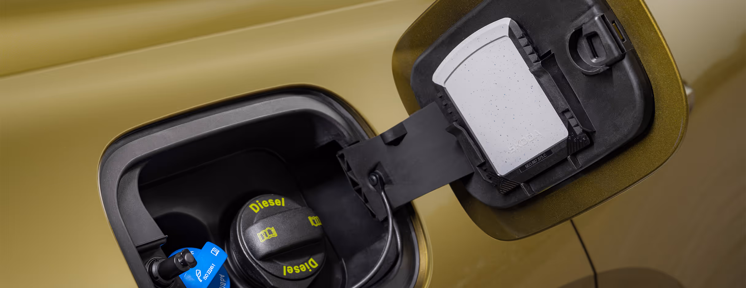 Eine Nahaufnahme zeigt die geöffnete Tankklappe eines Škoda Kodiaq in der Lackierung Gold-Bronze Metallic. Im Inneren sind der schwarze Tankdeckel für den Dieselkraftstoff und der danebenliegende blaue Deckel für den AdBlue-Zusatz zu sehen. An der Innenseite der Klappe ist der integrierte Eiskratzer als praktisches Detail untergebracht.