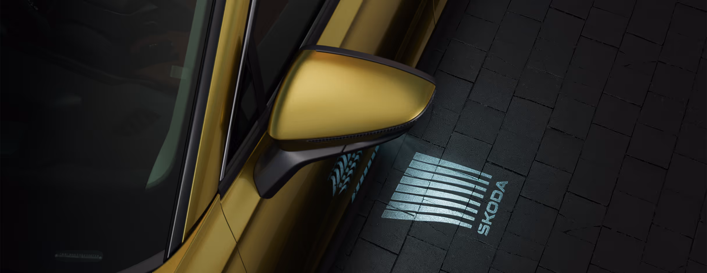Eine Nahaufnahme zeigt den Außenspiegel eines Škoda Kodiaq in der Lackierung Gold-Bronze Metallic bei Nacht. Aus dem Spiegel wird das Welcome-Logo, eine Grafik mit dem Škoda Schriftzug, auf den dunklen, gepflasterten Boden projiziert. Diese Einstiegsbeleuchtung erhellt den Bereich neben der Tür und begrüßt die Passagiere.