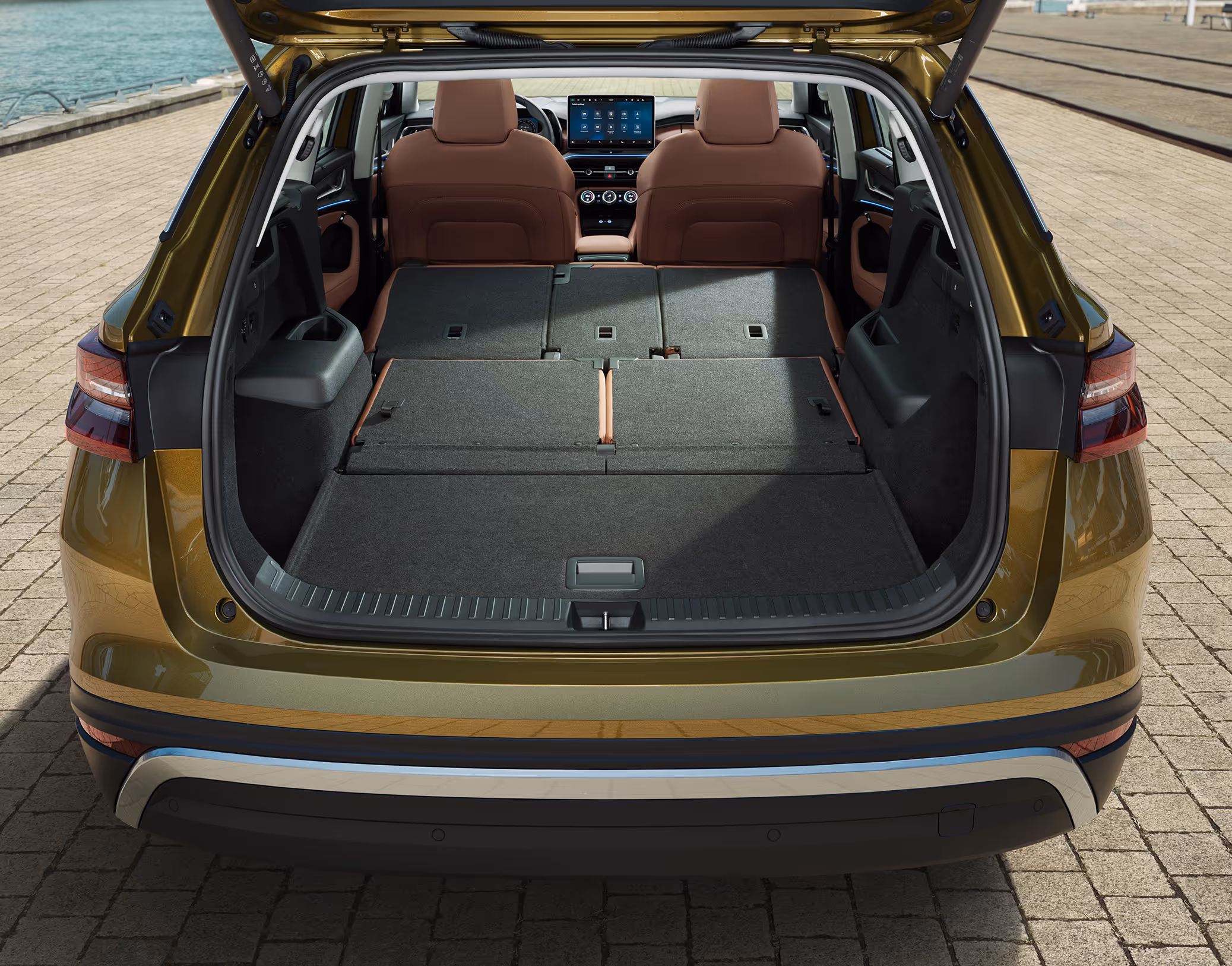 Eine Heckansicht zeigt den geöffneten Kofferraum eines Škoda Kodiaq in der Lackierung Gold-Bronze Metallic, der an einem Kai geparkt ist. Das Bild demonstriert eine praktische Stauraumlösung, bei der die Kofferraumabdeckung (Rollo) vertikal hinter den Rücksitzen verstaut ist. Im unteren Fach des variablen Ladebodens liegt ein farbenfroher, zusammengerollter Schlafsack.