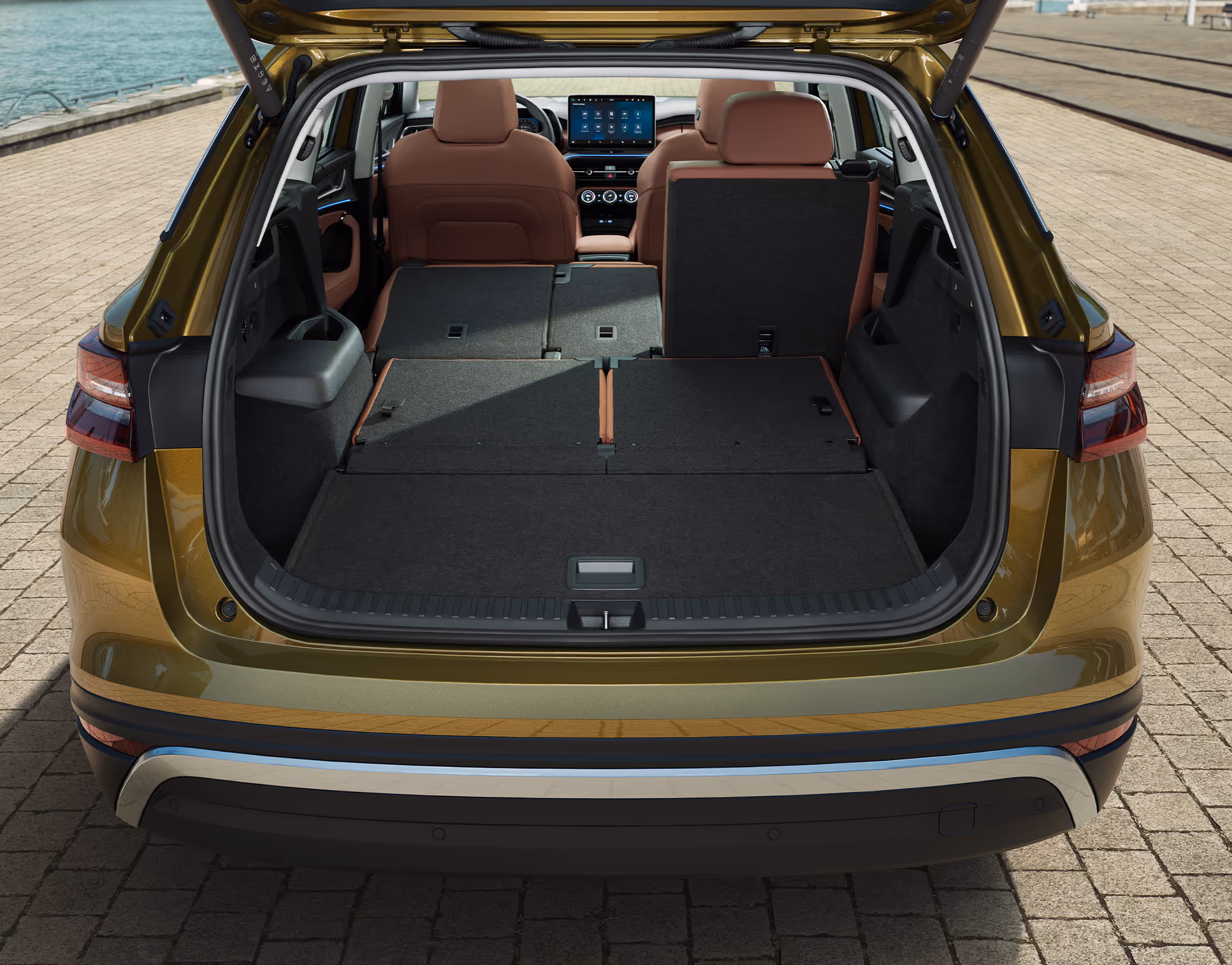 Eine Heckansicht zeigt den geöffneten Kofferraum eines Škoda Kodiaq in der Lackierung Gold-Bronze Metallic. Die zweite Sitzreihe ist teilweise, die dritte vollständig umgelegt, wodurch eine durchgehend ebene und sehr große Ladefläche entsteht. Das Fahrzeug parkt an einem Kai und im vorderen Bereich sind die aufrechten Vordersitze mit cognac-braunem Lederbezug zu sehen.