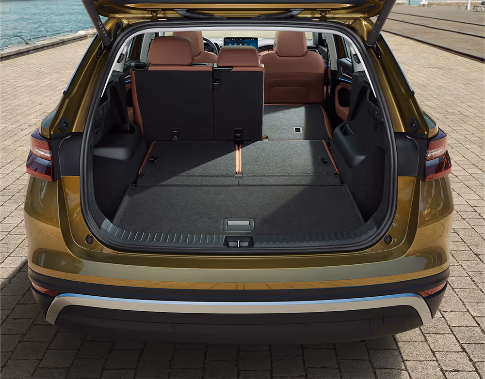 Eine Heckansicht zeigt den geöffneten Kofferraum eines Škoda Kodiaq in der Lackierung Gold-Bronze Metallic. Die zweite Sitzreihe ist teilweise, die dritte vollständig umgelegt, wodurch eine durchgehend ebene und sehr große Ladefläche entsteht. Das Fahrzeug parkt an einem Kai und im vorderen Bereich sind die aufrechten Vordersitze mit cognac-braunem Lederbezug zu sehen.