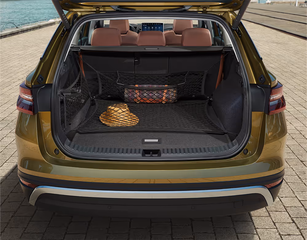 Eine Heckansicht zeigt den geöffneten Kofferraum eines Škoda Kodiaq in der Lackierung Gold-Bronze Metallic. Verschiedene schwarze Netze sichern die Ladung: Ein Bodennetz hält einen Strohhut fest, während ein vertikales Netz eine zusammengerollte Decke an ihrem Platz sichert. Durch den Kofferraum sind die cognac-braunen Ledersitze und die städtische Umgebung am Kai sichtbar.