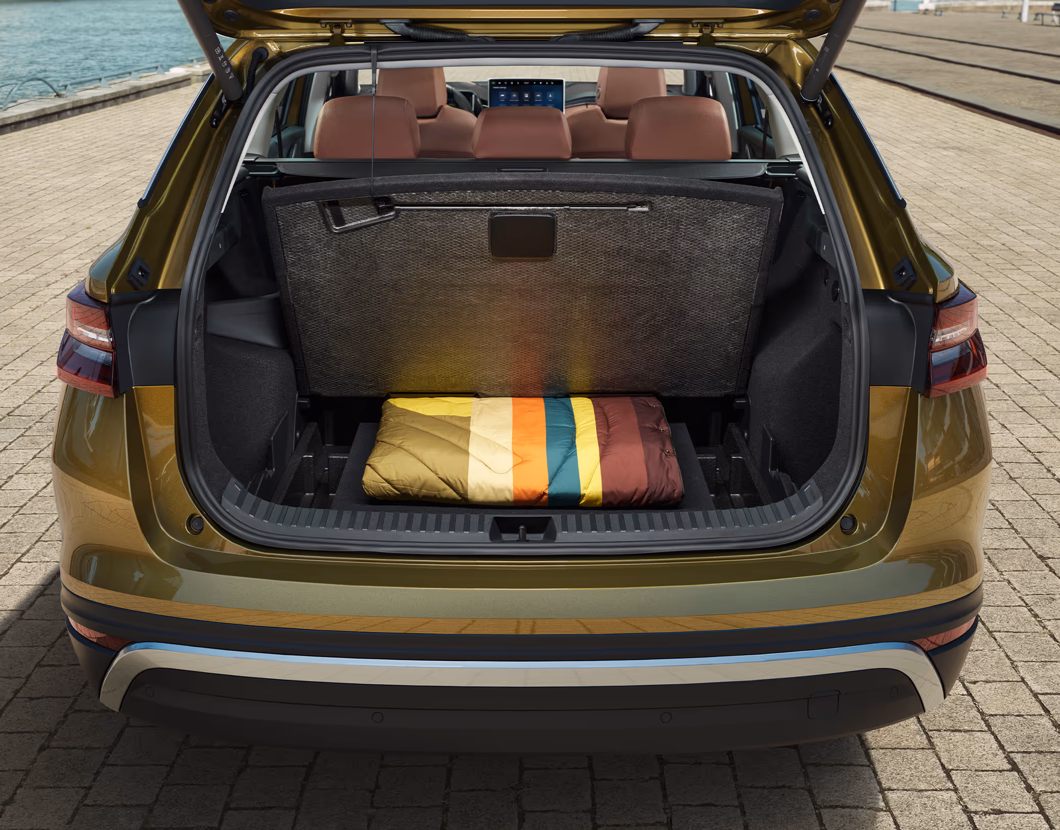 Eine Heckansicht zeigt den geöffneten Kofferraum eines Škoda Kodiaq in der Lackierung Gold-Bronze Metallic, der an einem Kai geparkt ist. Das Bild demonstriert eine praktische Stauraumlösung, bei der die Kofferraumabdeckung (Rollo) vertikal hinter den Rücksitzen verstaut ist. Im unteren Fach des variablen Ladebodens liegt ein farbenfroher, zusammengerollter Schlafsack.