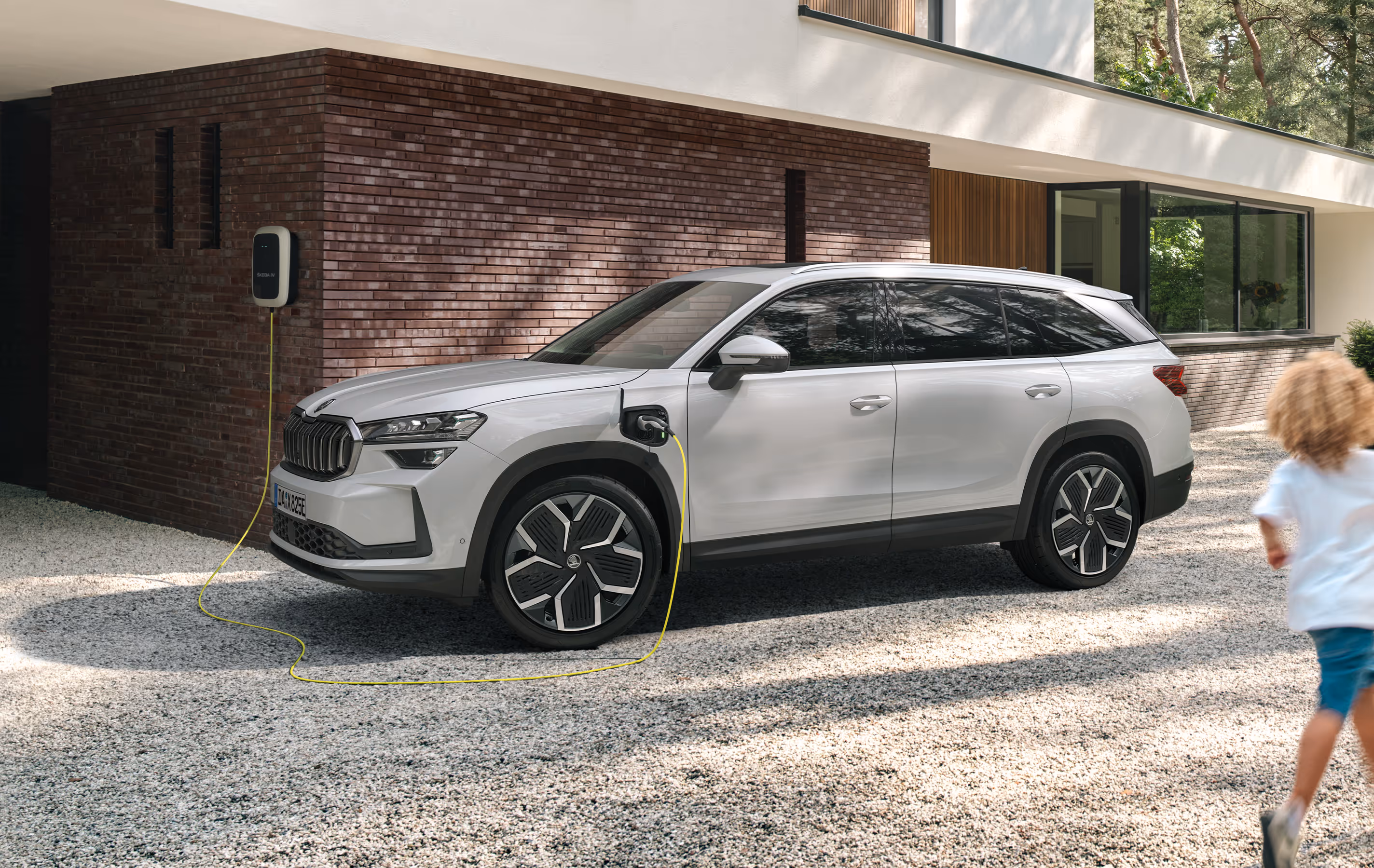 Ein Škoda Kodiaq iV Plug-in-Hybrid mit der Lackierung Moon-Weiß Perleffekt lädt an einer Wallbox in der Kieseinfahrt eines modernen Hauses. Das Fahrzeug ist über ein Ladekabel mit der Ladestation verbunden, während im Vordergrund ein kleines Kind auf einen Ball zuläuft. Rechts unten steht auf einer dunkelgrün abgesetzten Fläche "Kodiaq iV" geschrieben.