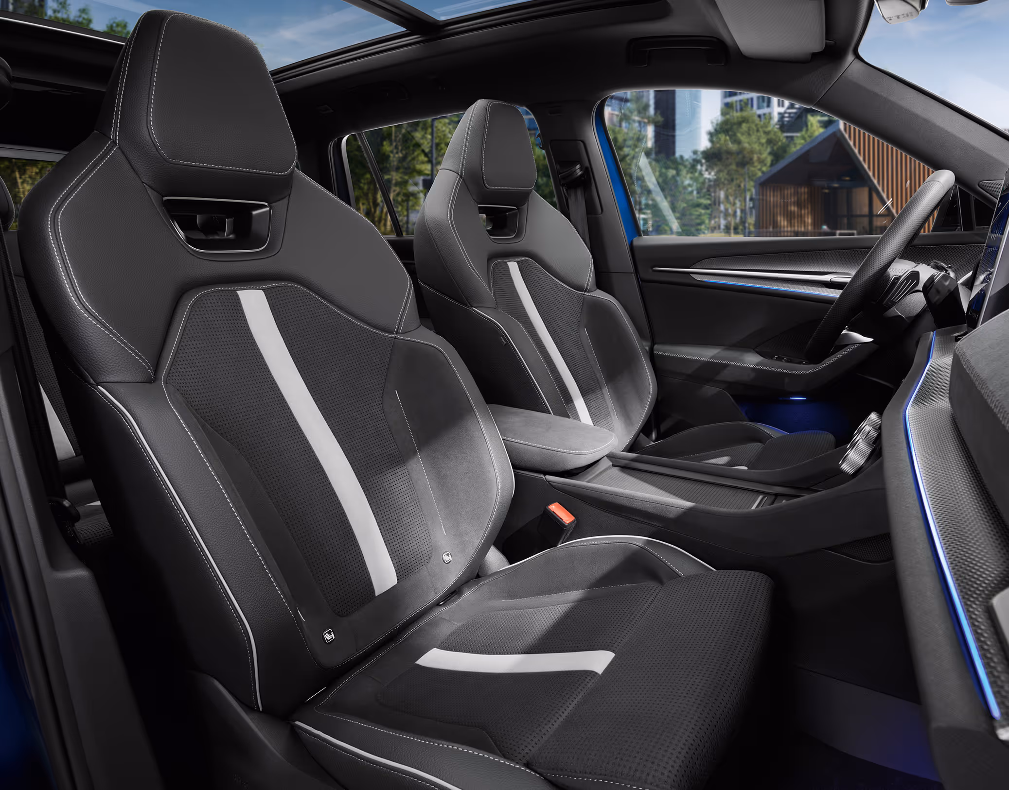 Eine Innenansicht des Škoda Kodiaq Sportline zeigt die vorderen Sportsitze mit integrierten Kopfstützen. Die Sitze der Design Selection Sportline sind mit einer Kombination aus schwarzem, perforiertem Microfaser-Stoff und Kunstleder bezogen und weiß-gräulichen Zierstreifen sowie Kontrastnähte auf. Durch das große Panoramaschiebedach fällt Tageslicht in den Innenraum und gibt den Blick auf eine von Bäumen gesäumte, städtische Umgebung frei.