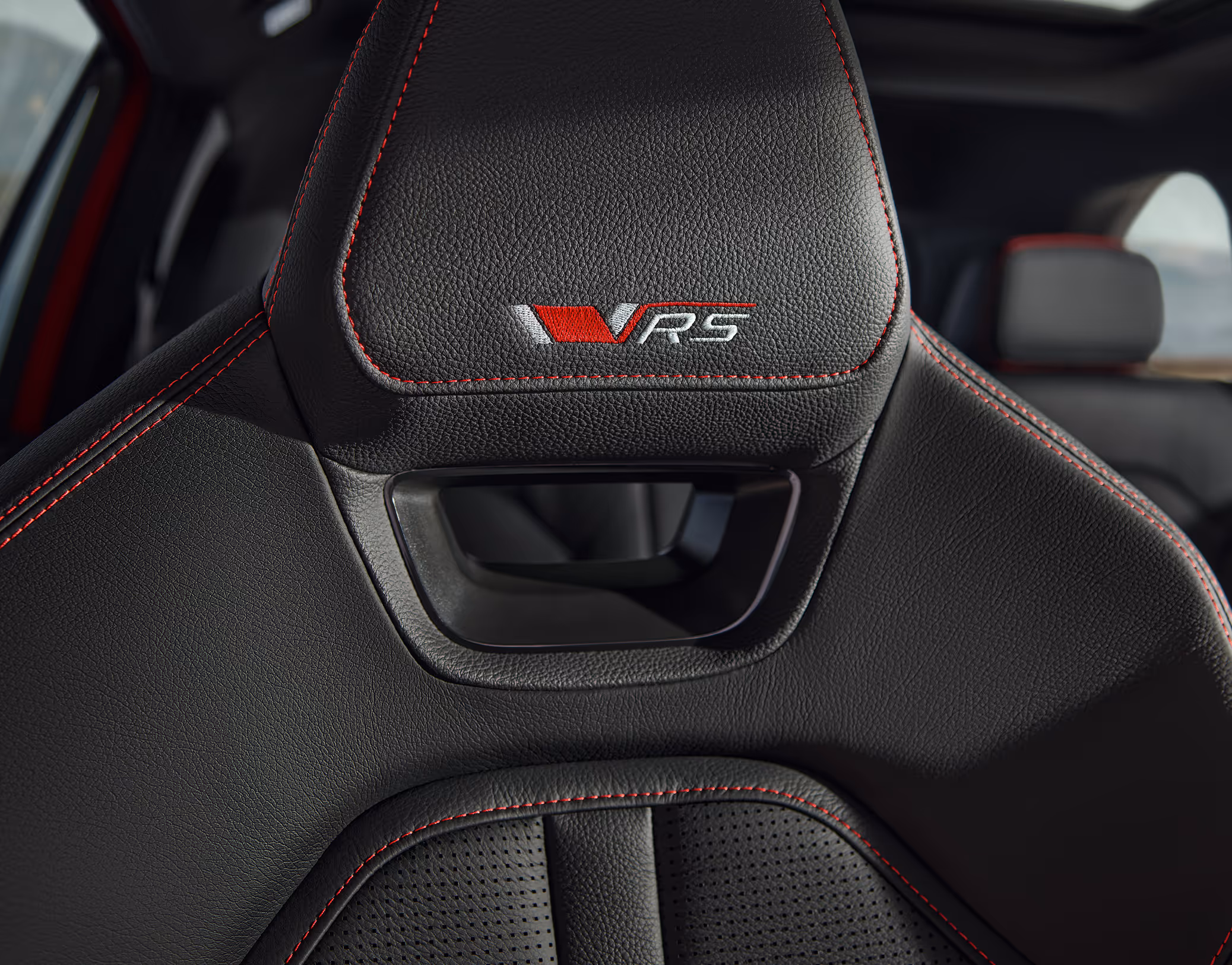 Eine Nahaufnahme des Sportsitzes im Škoda Kodiaq RS zeigt die integrierte Kopfstütze. Das eingestickte RS-Logo und die markanten roten Kontrastnähte auf dem schwarzen, perforierten Lederbezug heben den dynamischen Charakter der Design Selection RS Lounge hervor. Im unscharfen Hintergrund sind das Panoramaschiebedach und die Landschaft außerhalb des Fahrzeugs zu erkennen.