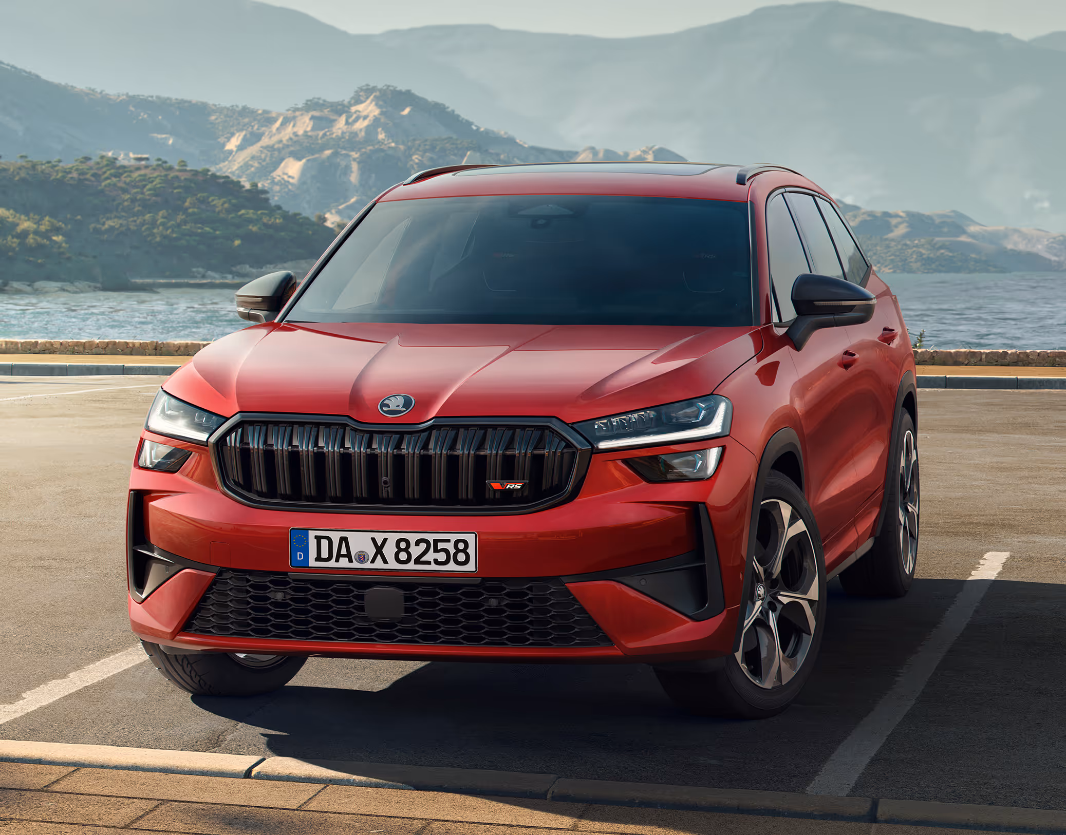 Ein Škoda Kodiaq RS in der Lackierung Velvet-Rot Metallic parkt auf einem Parkplatz vor einer malerischen Küsten- und Berglandschaft. Die frontale Dreiviertelansicht hebt die sportlichen Designmerkmale des RS-Modells hervor, darunter der glänzend schwarze Kühlergrill mit RS-Logo und die schwarzen Außenspiegelkappen.